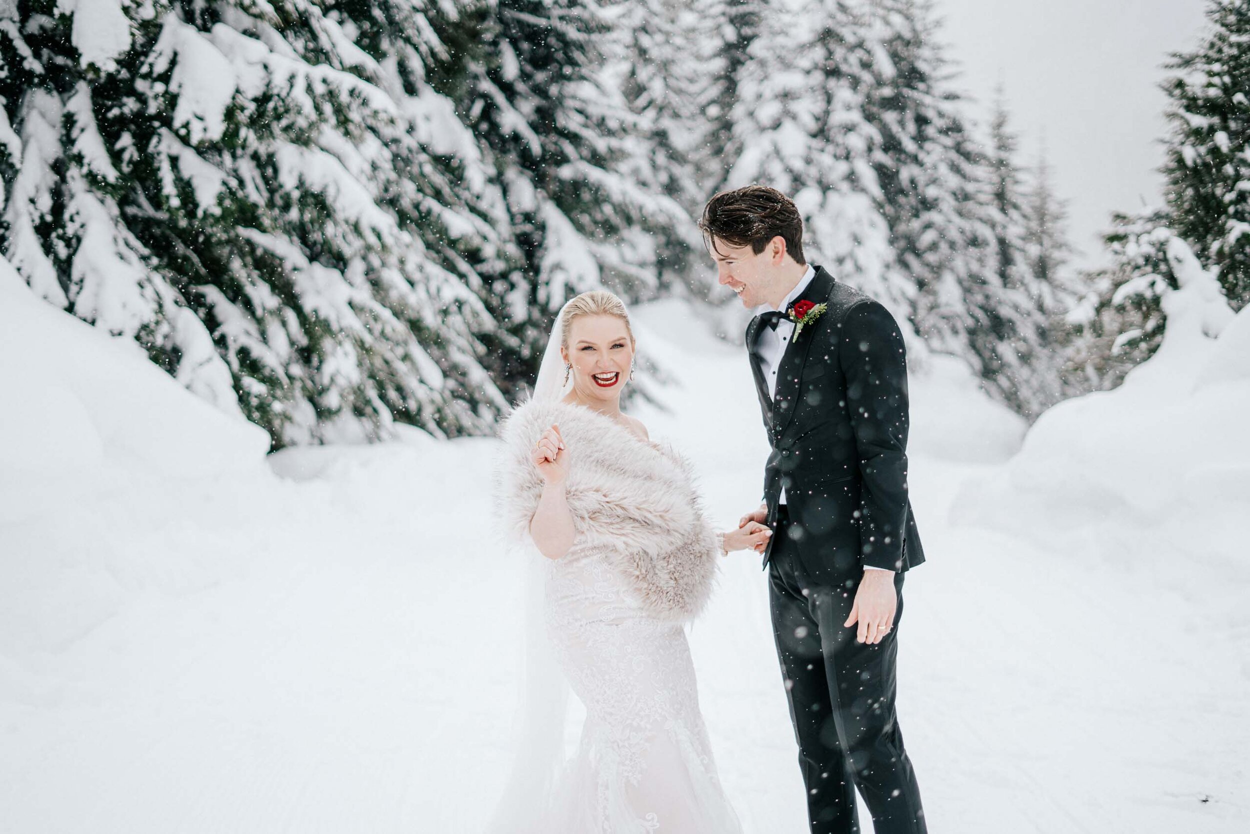 A bride smiles at the camera in the snow as her groom holds her hand and the snow falls.