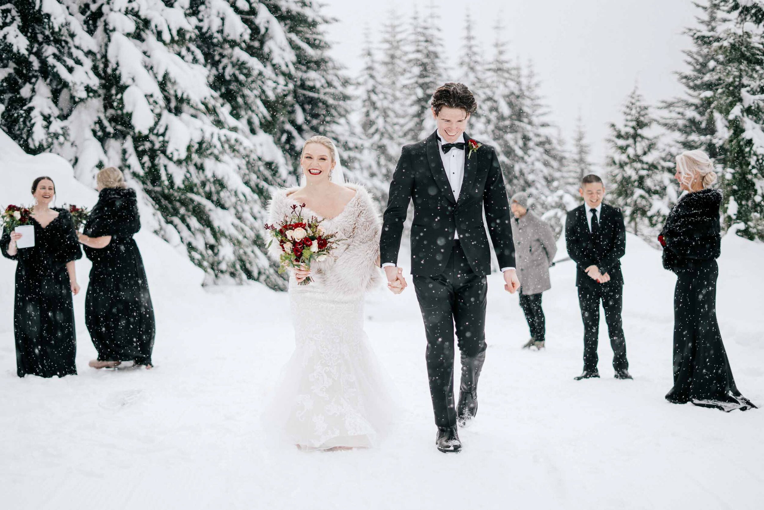 A bride and groom hold hands as they walk through the falling snow during their outdoor Whistler wedding. 