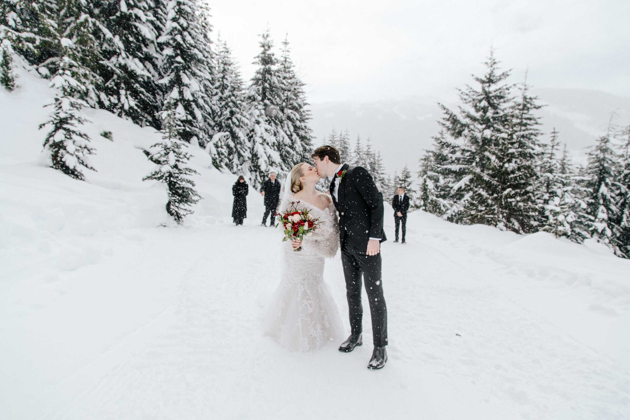 A bride in a fur stole and lace dress kisses her groom in the snow.