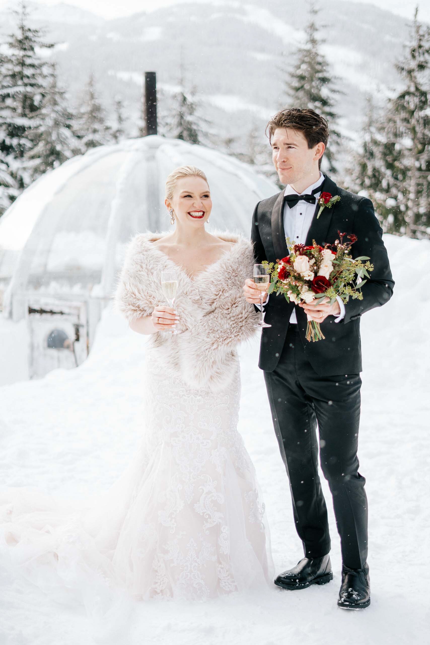 A  chilly bride covers herself with an umbrella as the snow falls and her groom holds her umbrella for her.