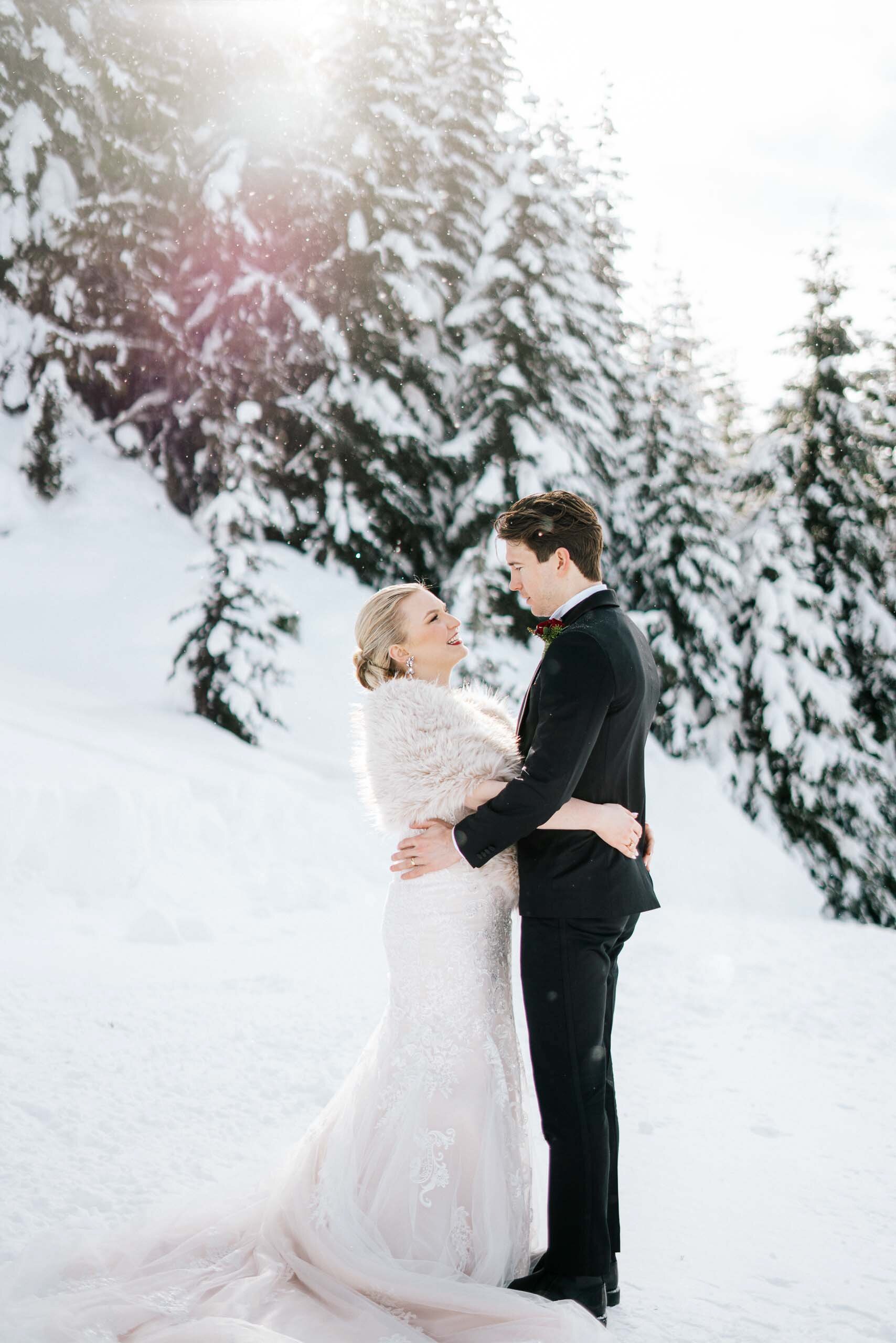 A bride in a fur stole holds her groom surrounded by snowy evergreens at their Whistler BC Wedding. 