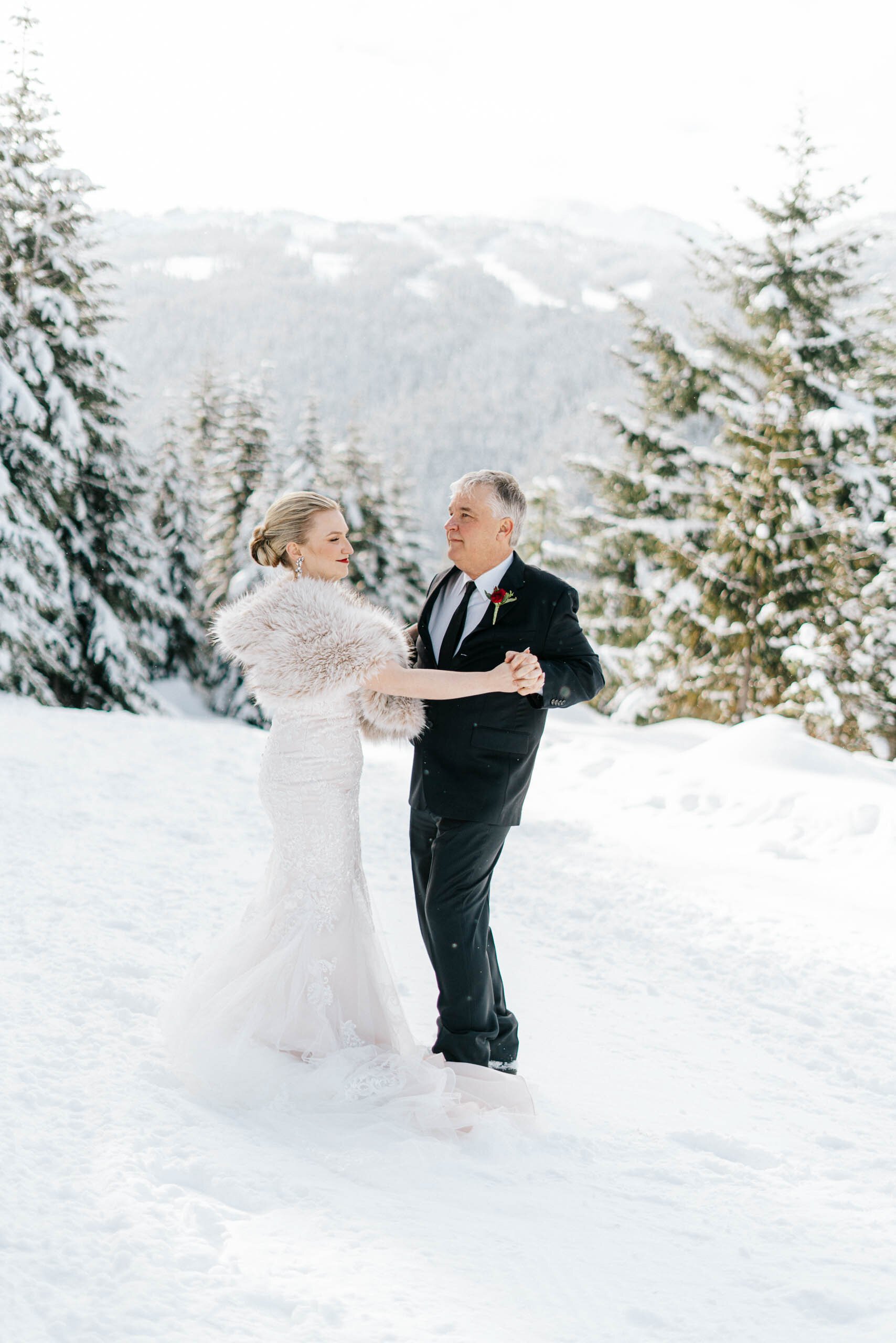 A Bride in lace dress and fur stole dances with her father in the snow in front of beautiful evergreen trees. 