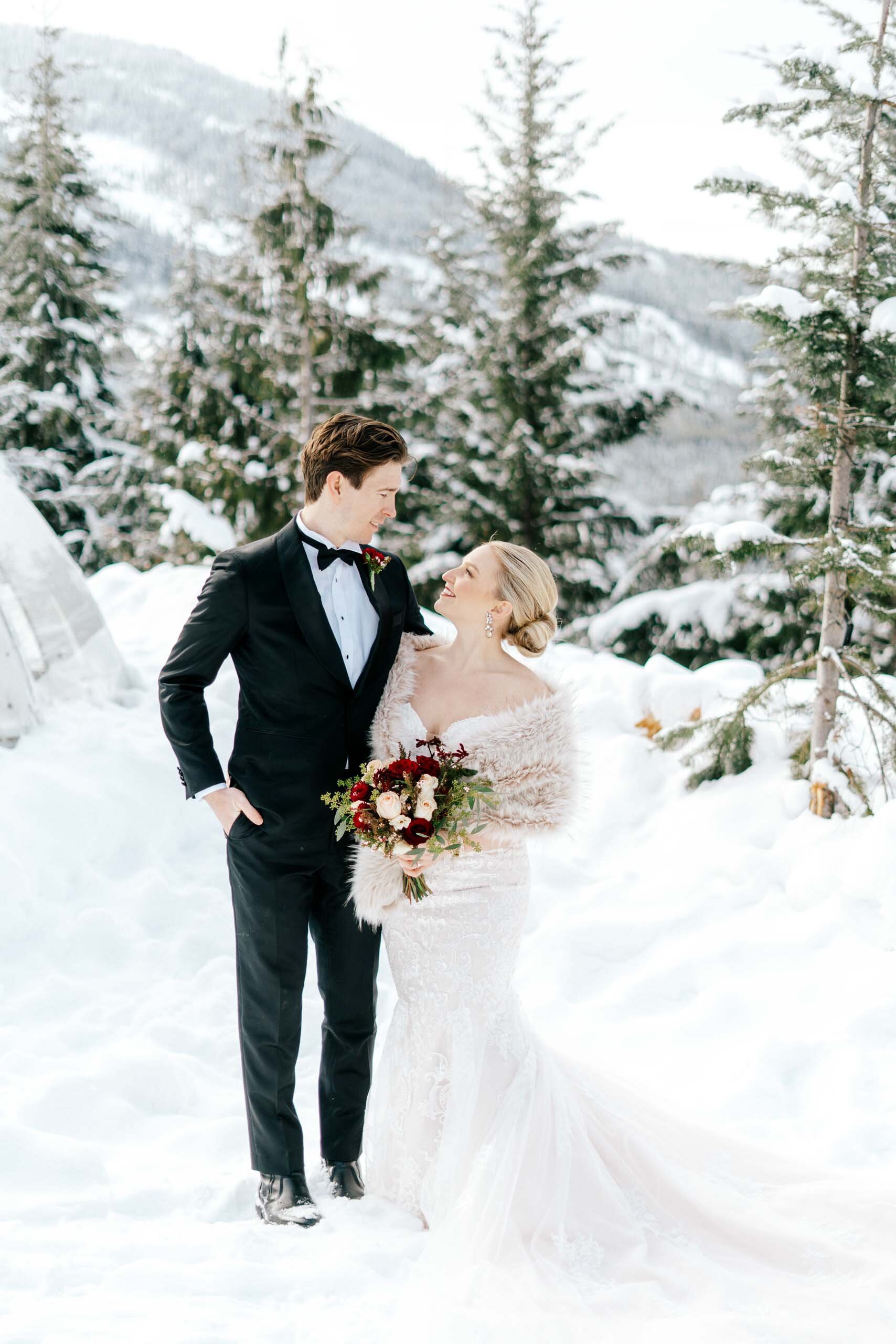 A groom looks adoringly at his bride as they stand in the snow. 