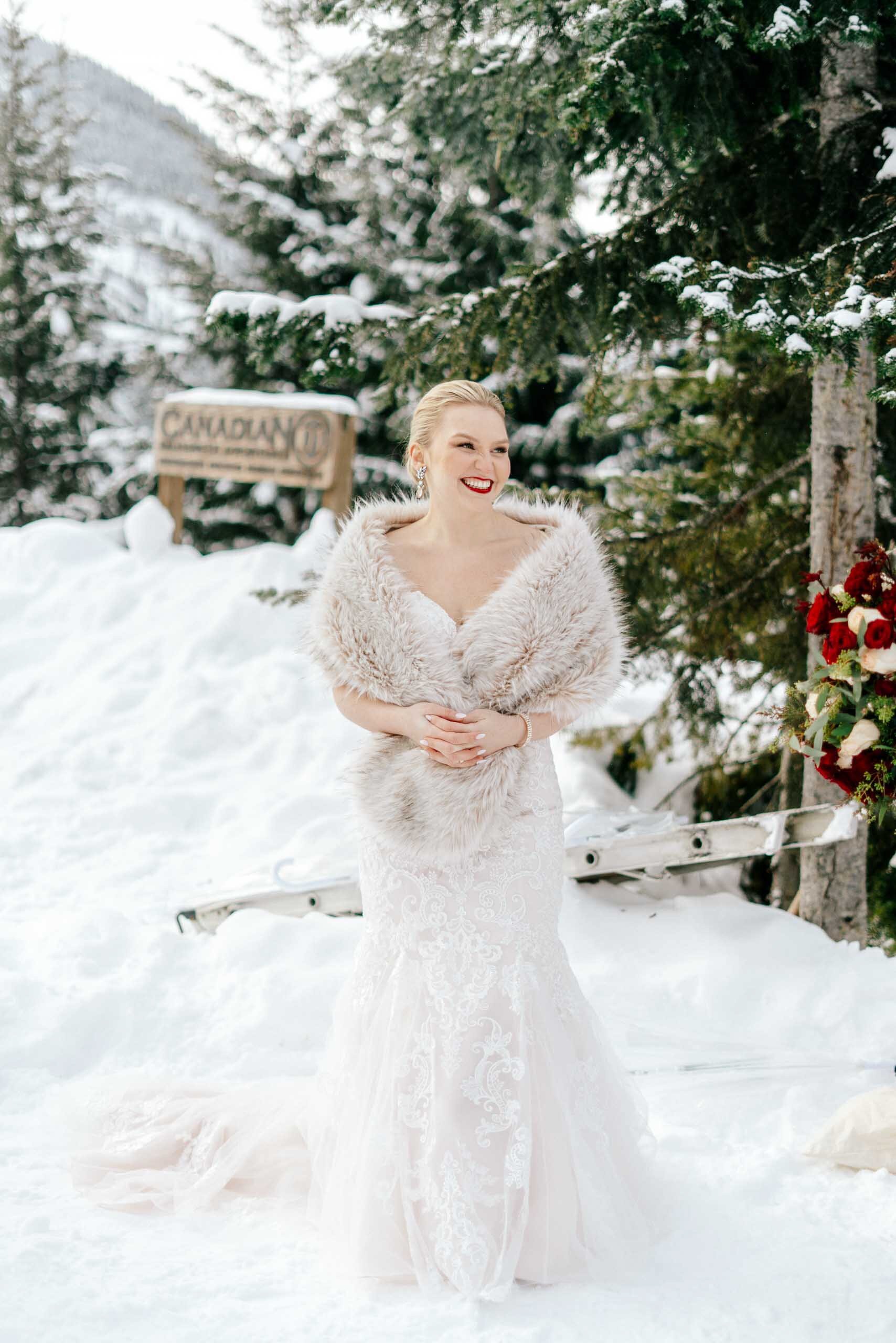 A bride wraps her soft fur stole around herself as she smiles in the snow on her wedding day.