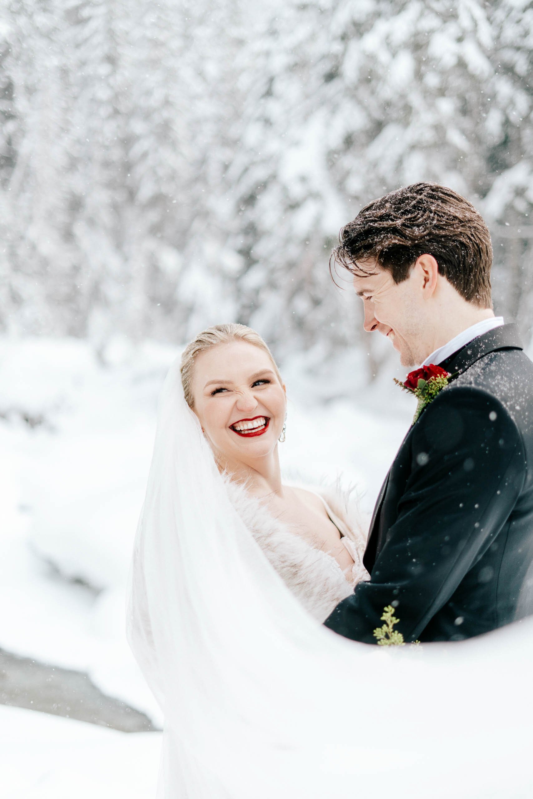 A blonde bride with bright red lip stick smiles at the camera as her veil swoops around her and her groom laughs.