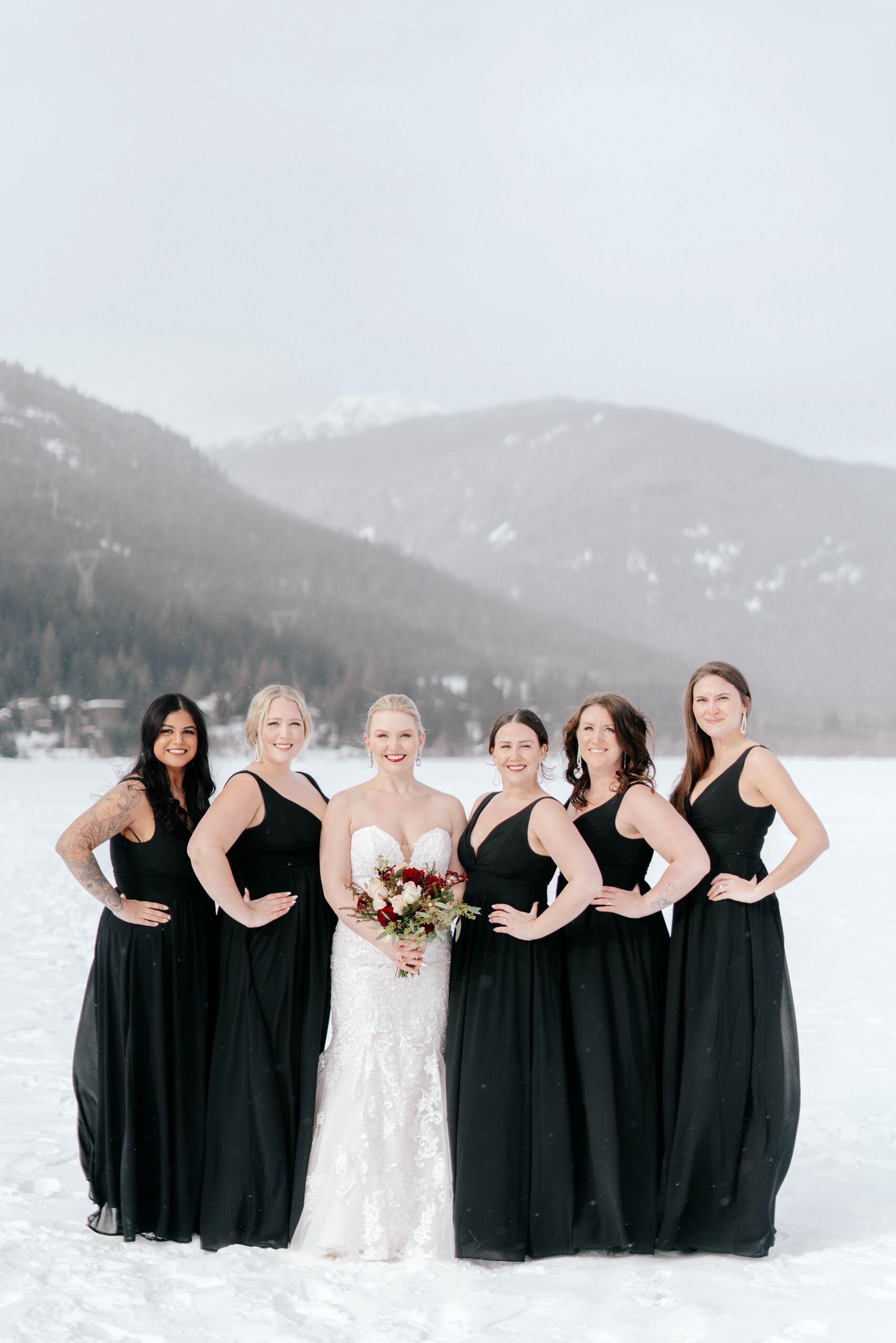 A bridal party in elegant black evening gowns stand with the bride in the snowy mountains of Whistler, BC.