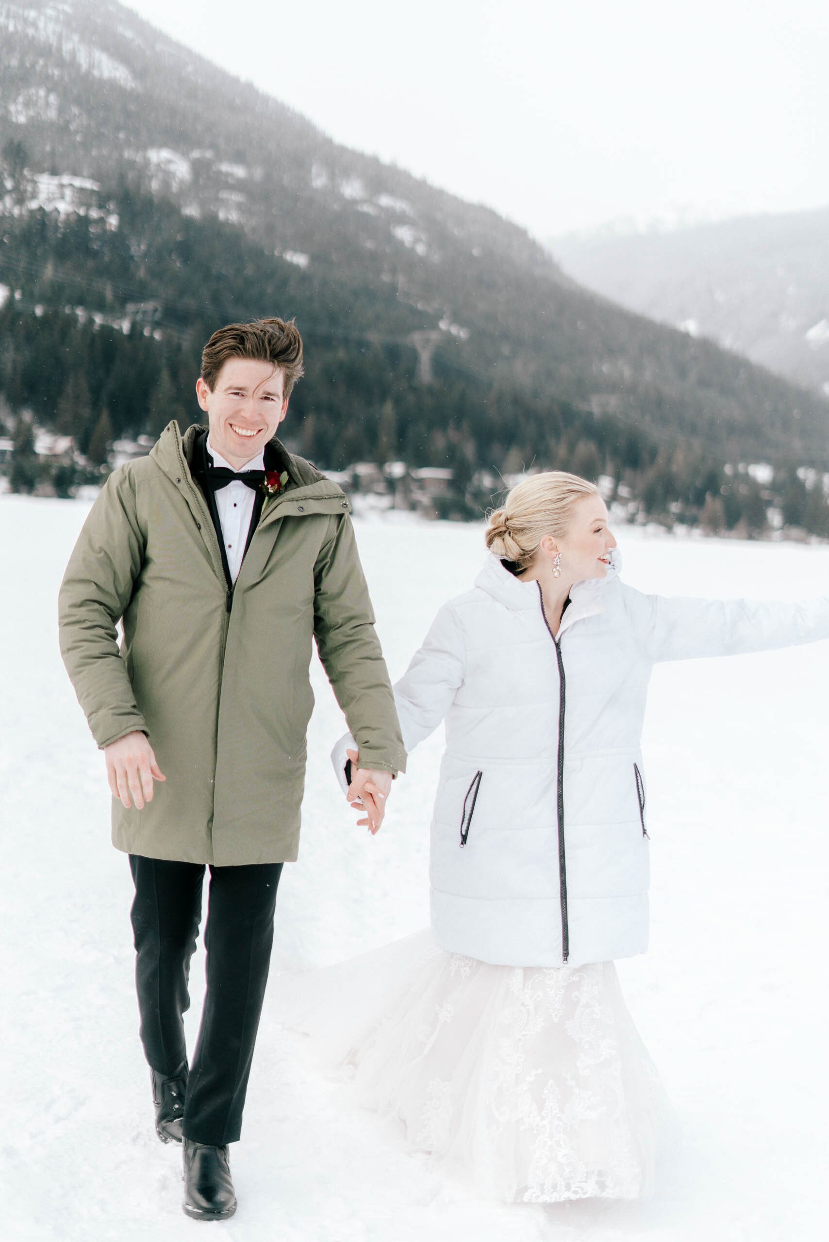 A bride and groom walk through the snow while wearing winter jackets over their wedding attire. 