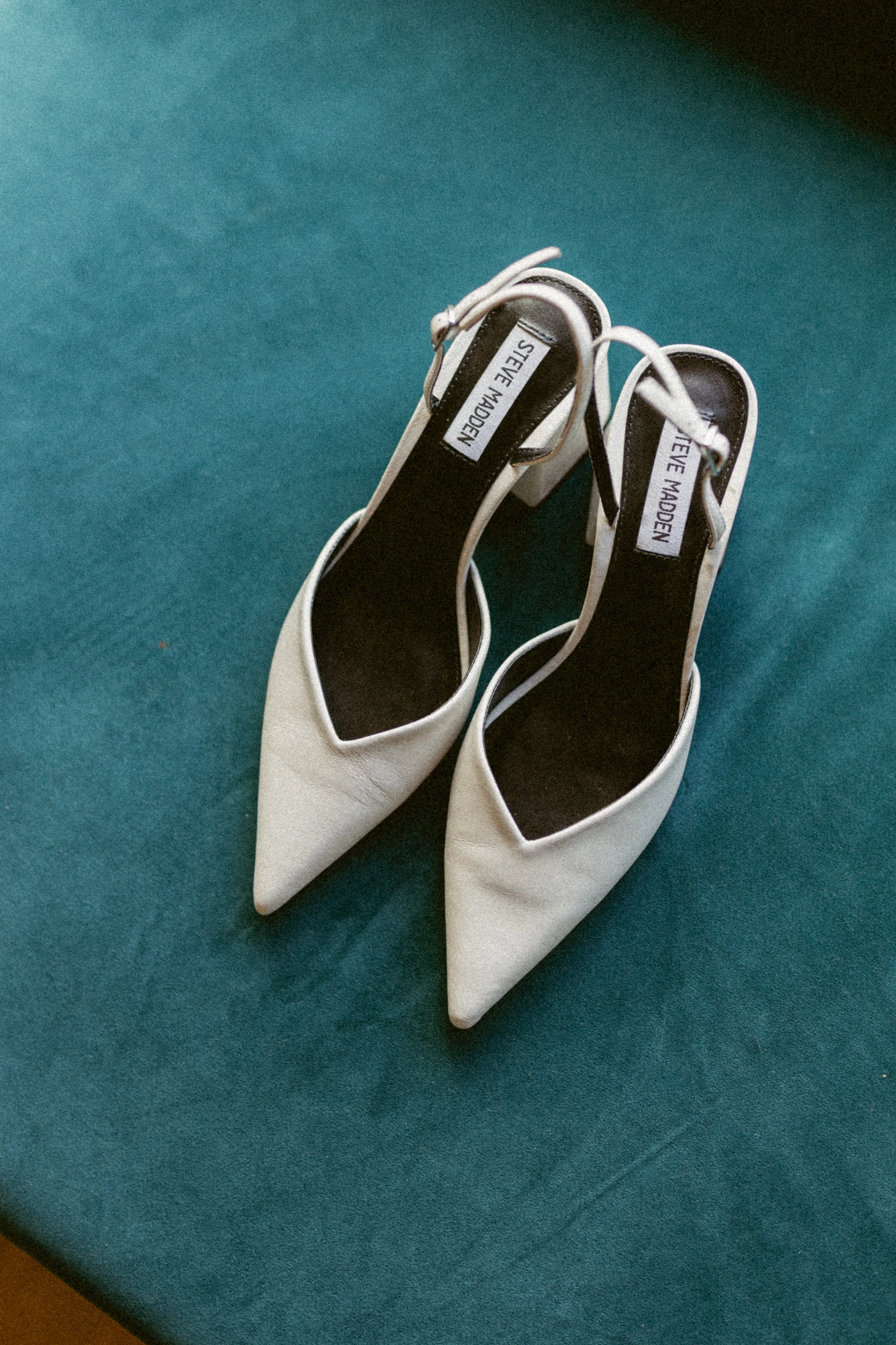 Editorial detail shot of white pointed-toe bridal heels placed on a teal velvet surface.