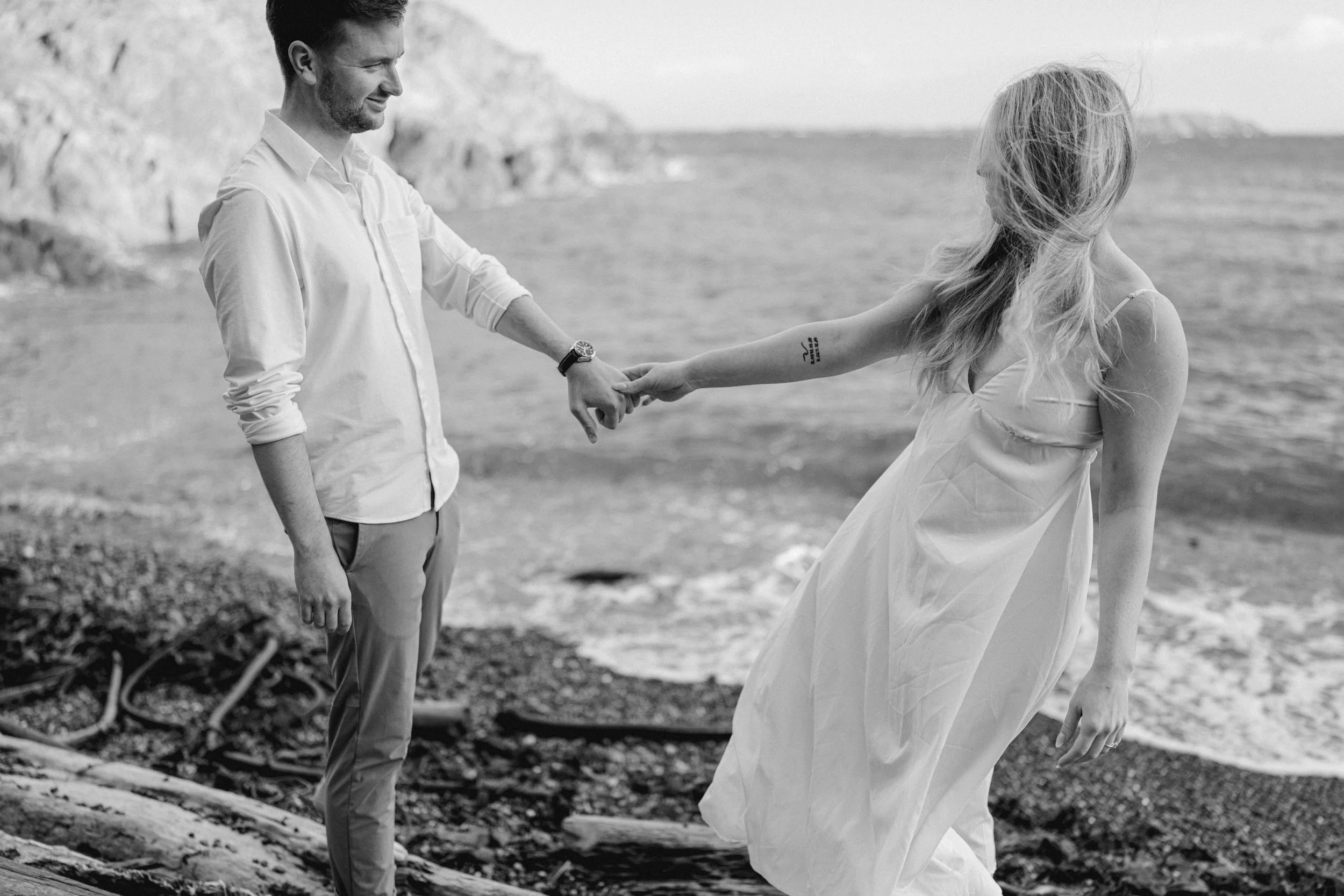 Black and white engagement photo of a couple holding hands along the rocky beach at Dallas Road.