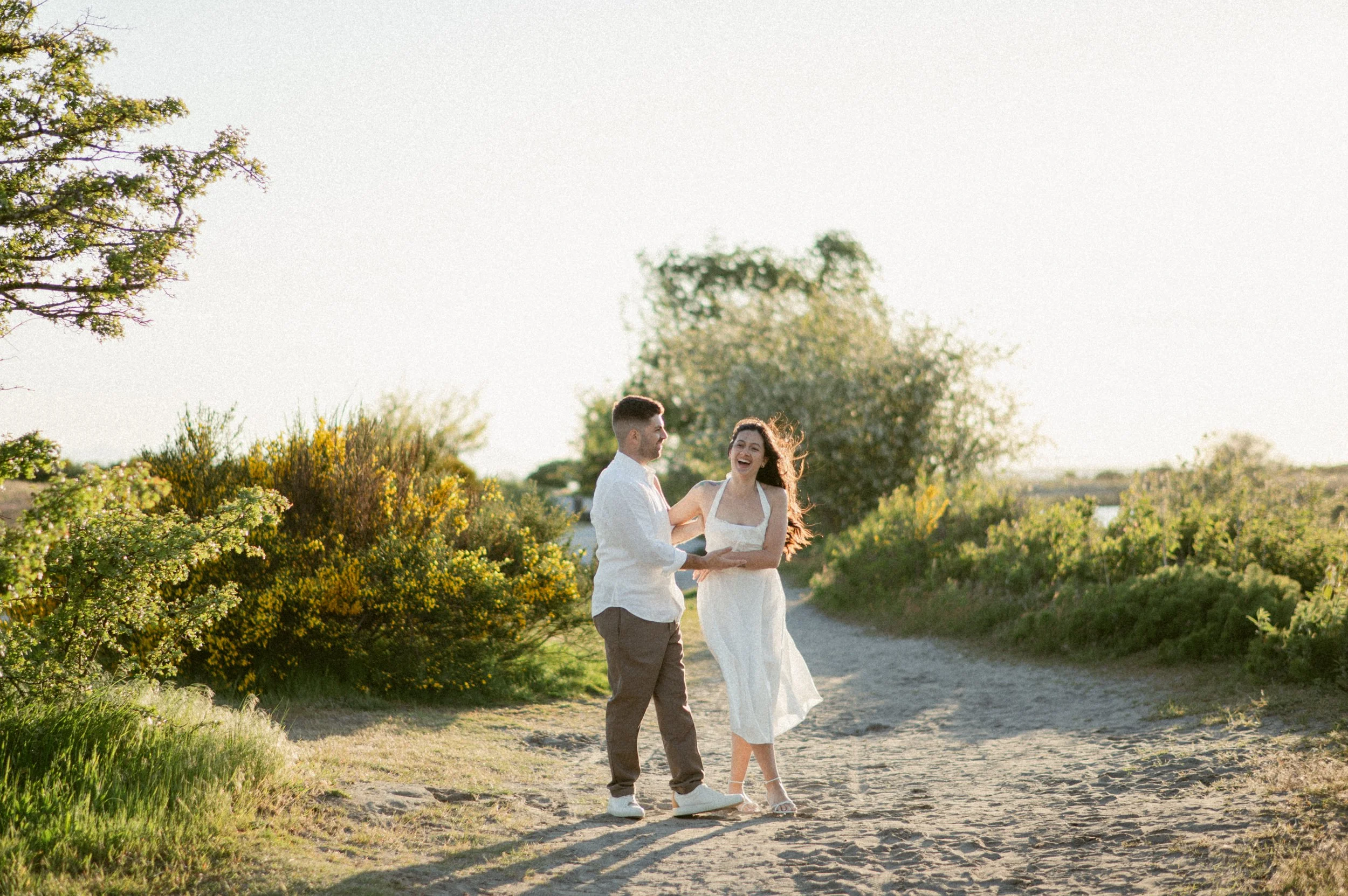 Engaged couple laughing and holding hands on a sandy path at Gary Point Park during golden hour.
