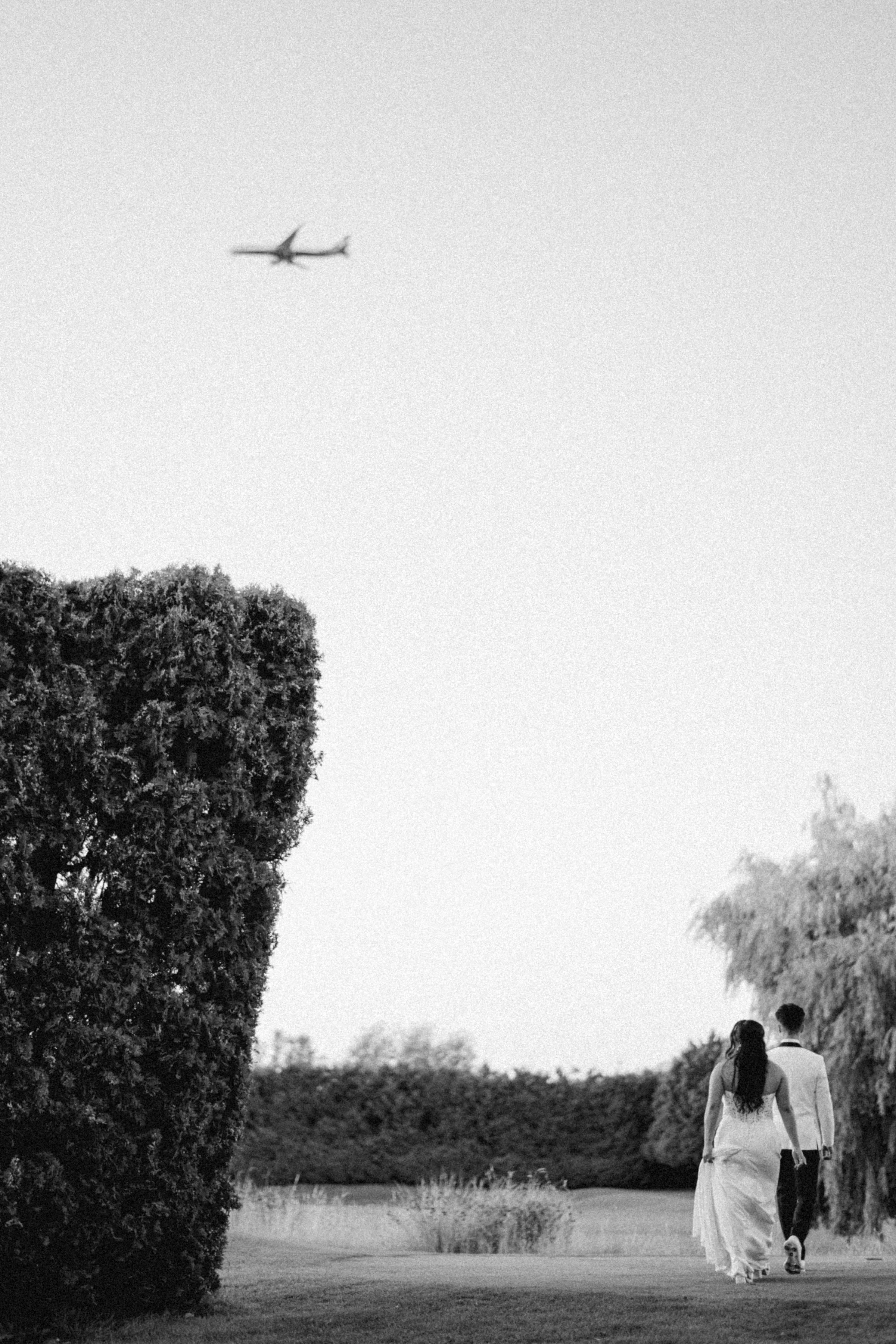Black and white photograph of a couple walking together across an open field with an airplane in the sky above them.