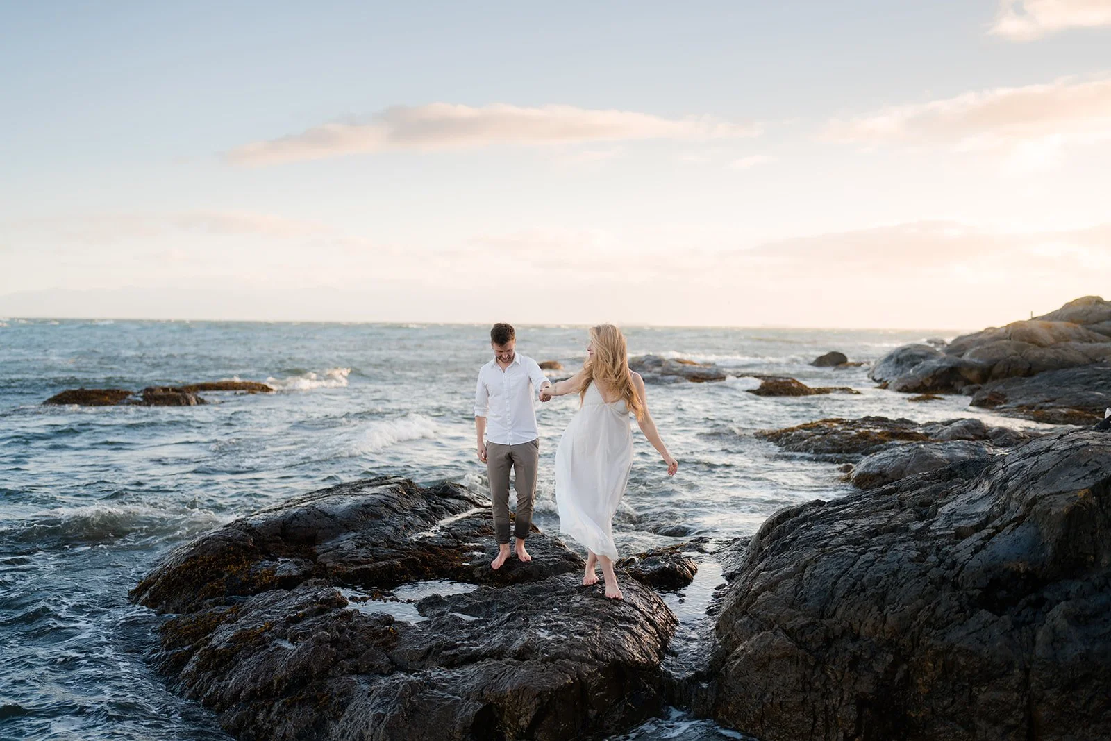 Engaged couple walking barefoot across rocky shoreline at sunset on Dallas Road in Victoria.