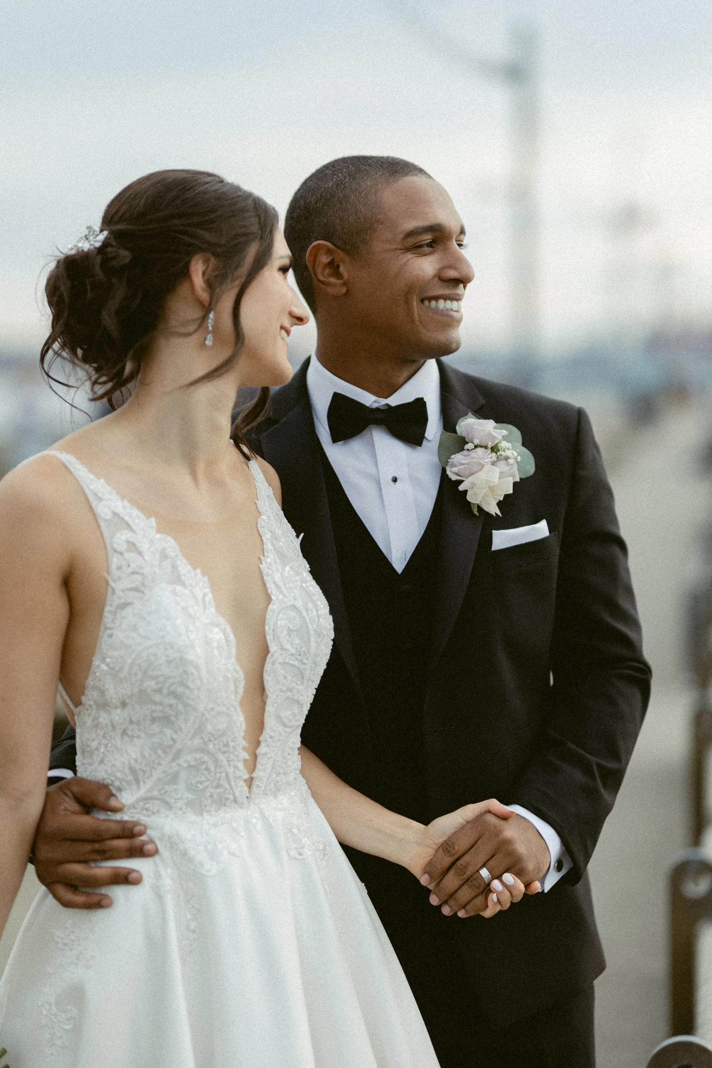 Bride and groom stand hand in hand near the water, smiling as they look into the distance.