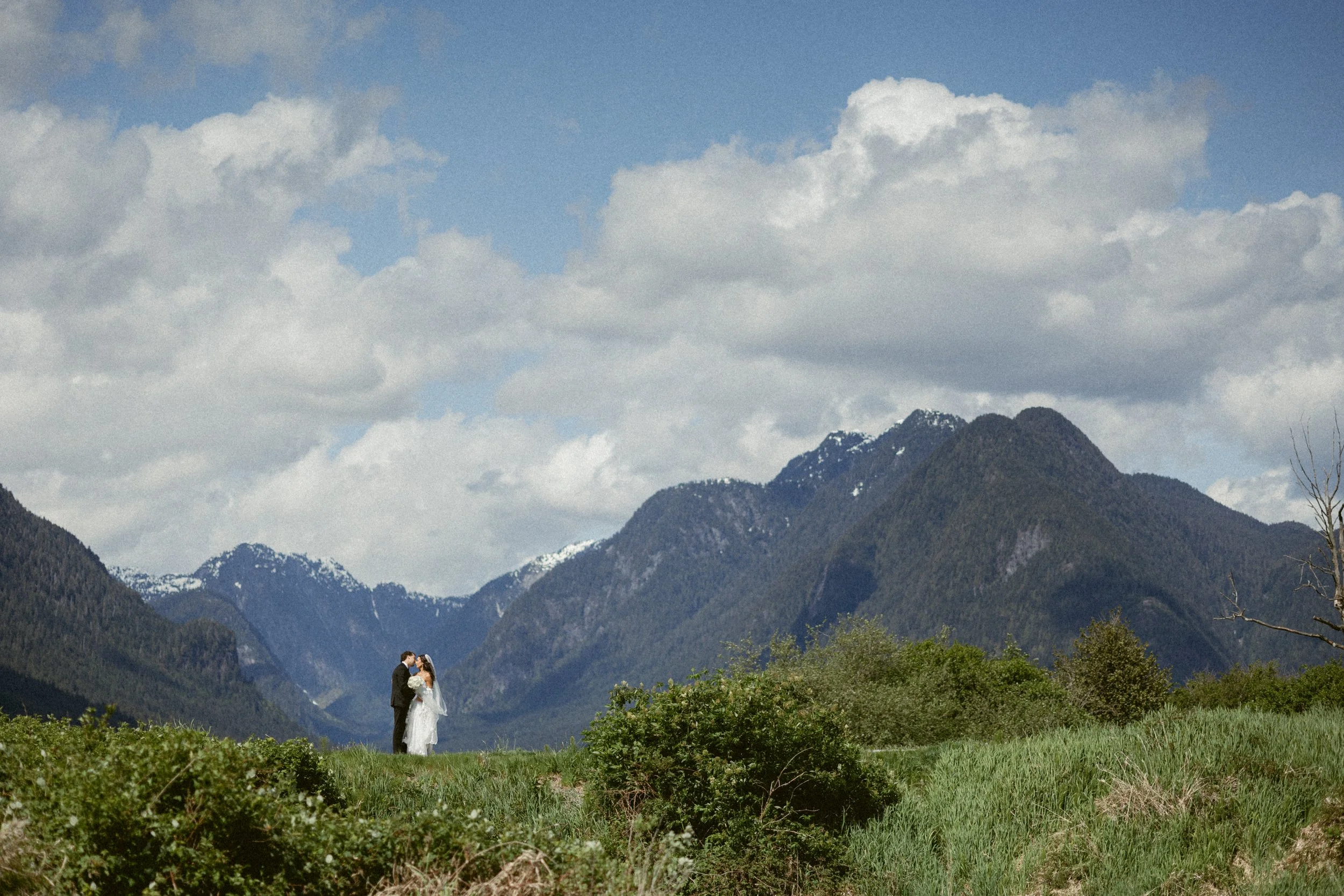 Bride and groom stand together in a wide mountain landscape, sharing a quiet kiss at Pitt Lake.