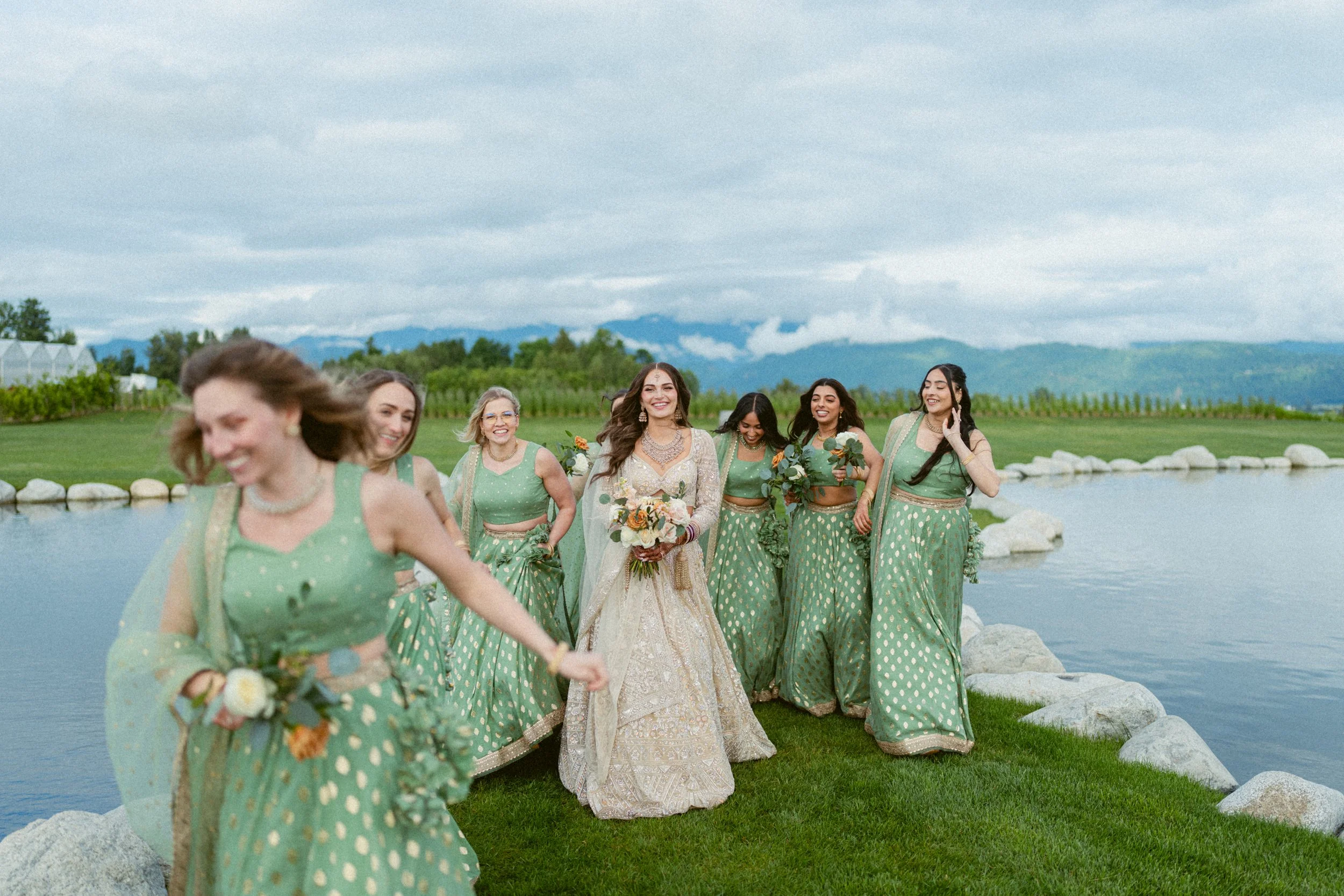 Bride walking with bridesmaids in coordinated green outfits beside a pond at Mann Farms.