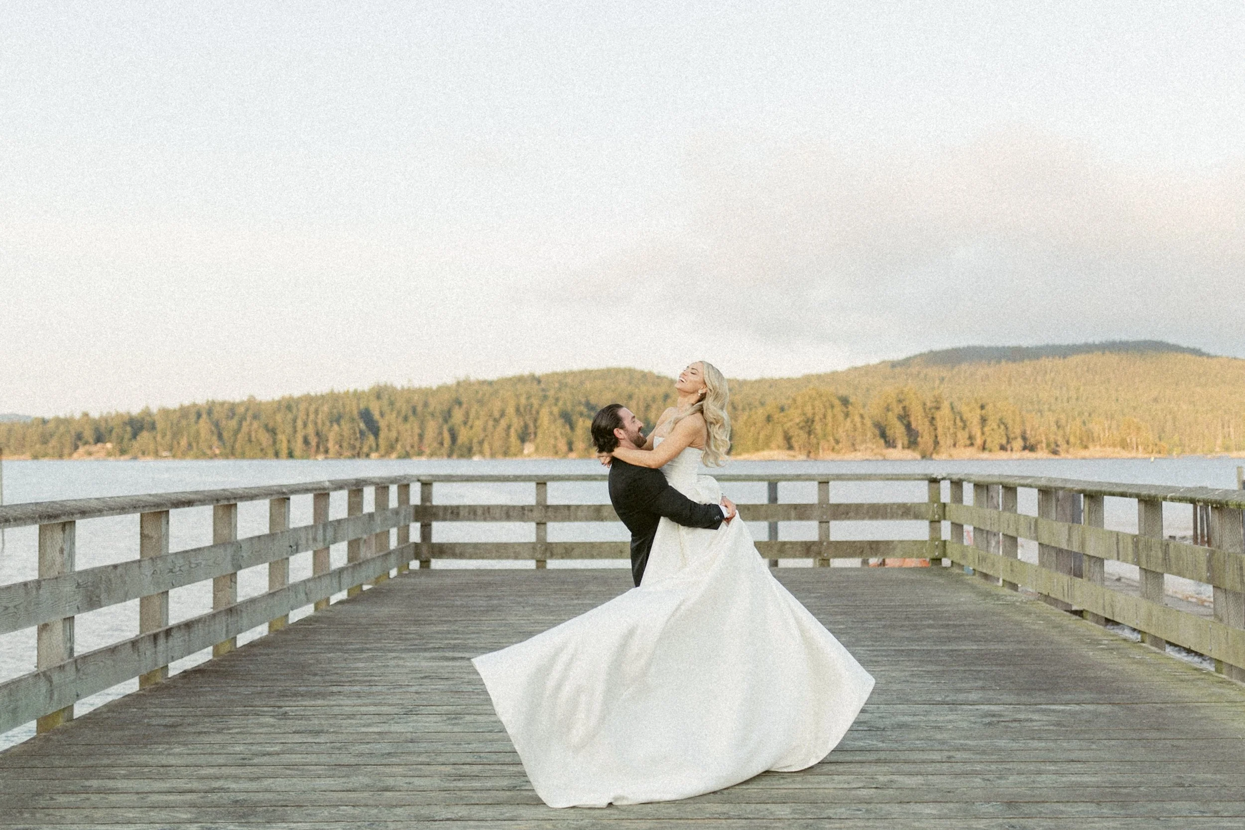 Bride and groom laughing as the groom lifts the bride on a wooden pier overlooking the ocean near Prestige Oceanfront Resort in Sooke.