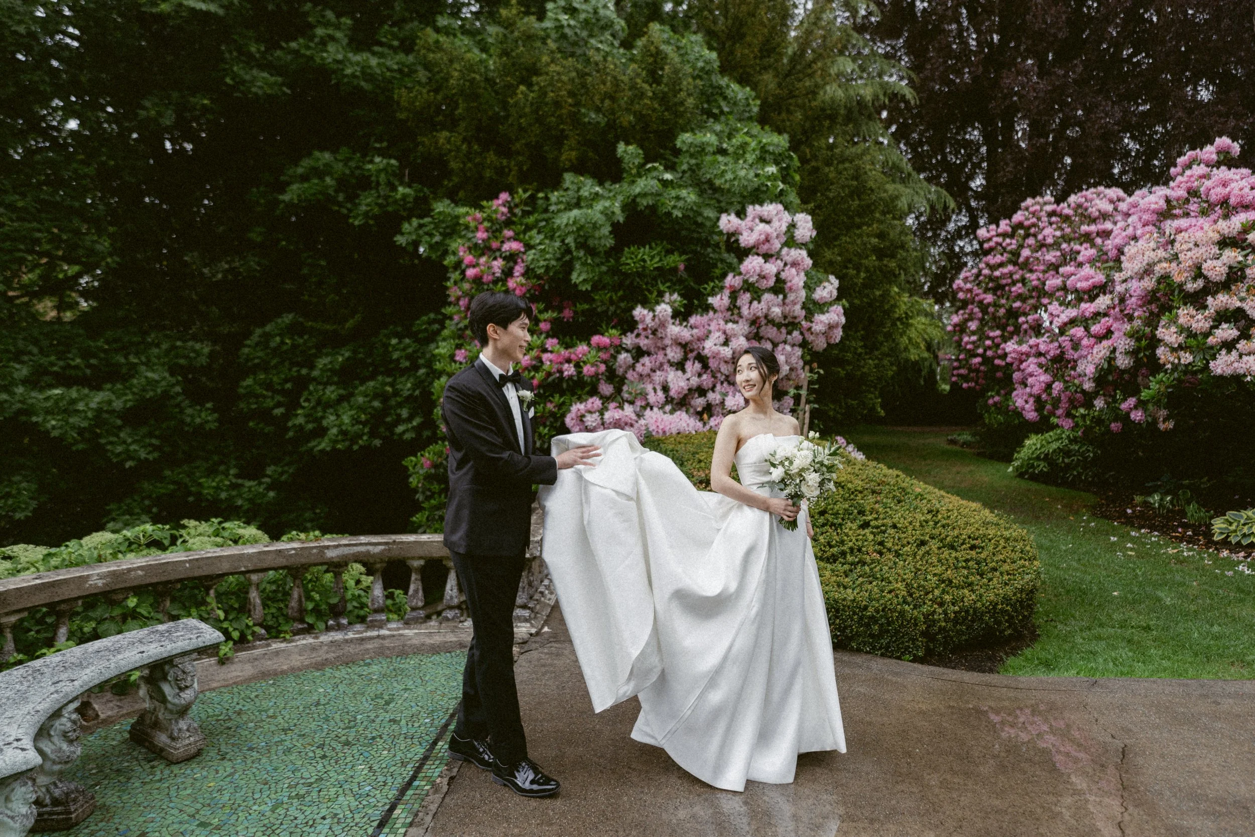 Bride and groom walking together through a garden framed by pink blossoms.