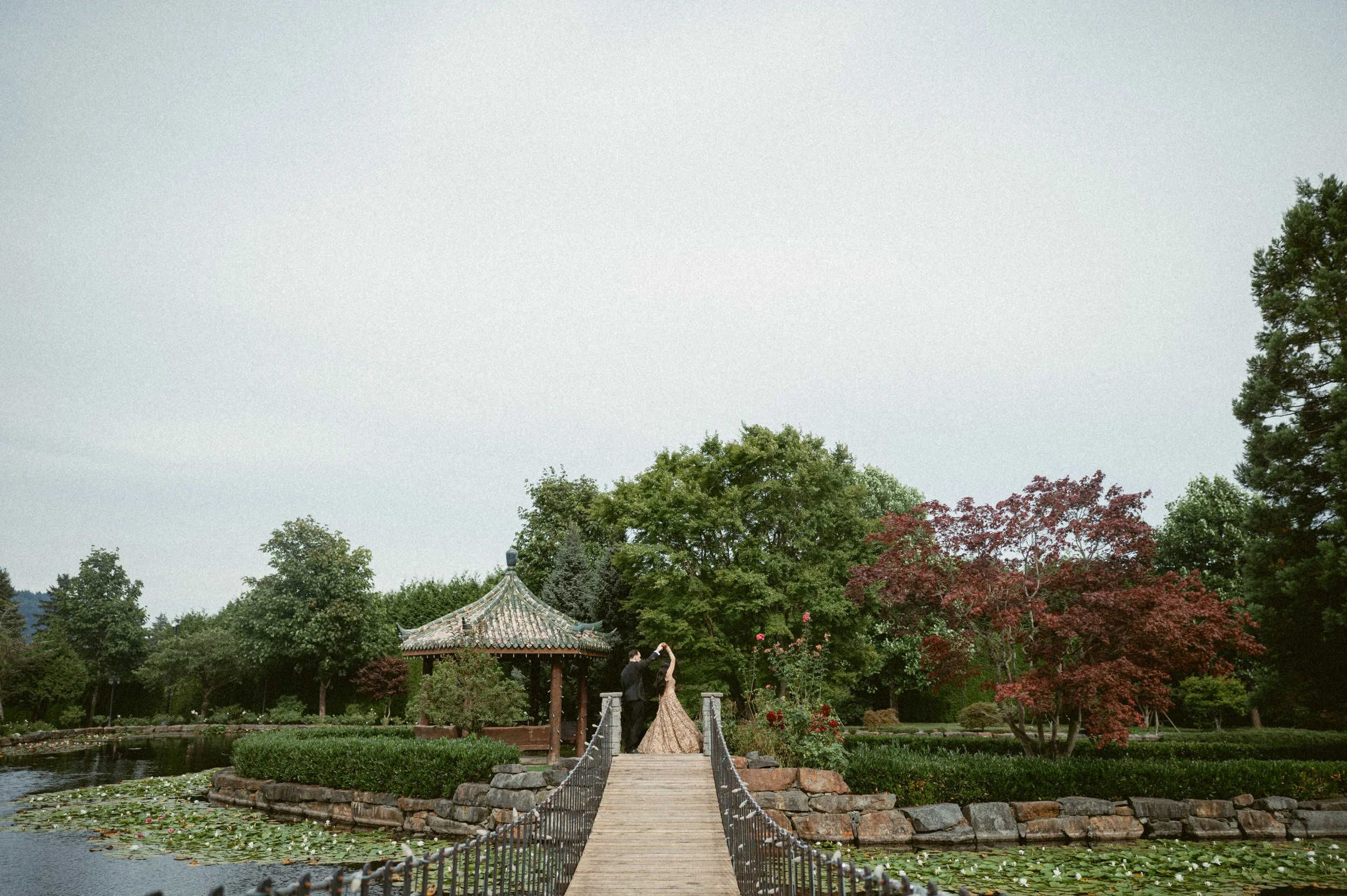 Wide shot of a bride and groom dancing on a bridge in a landscaped garden with trees and water surrounding them.