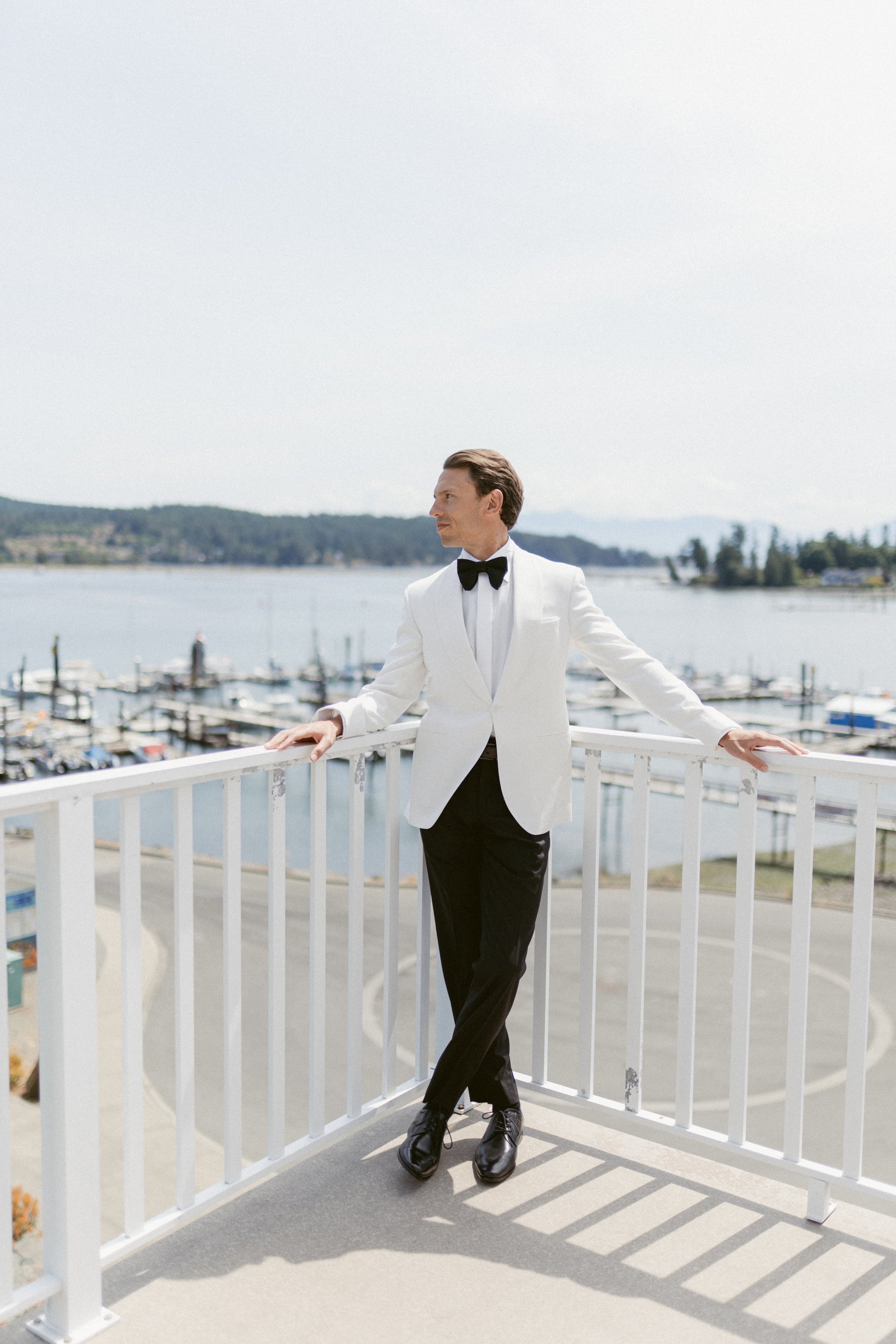 Groom leaning on white railing with ocean in the background.