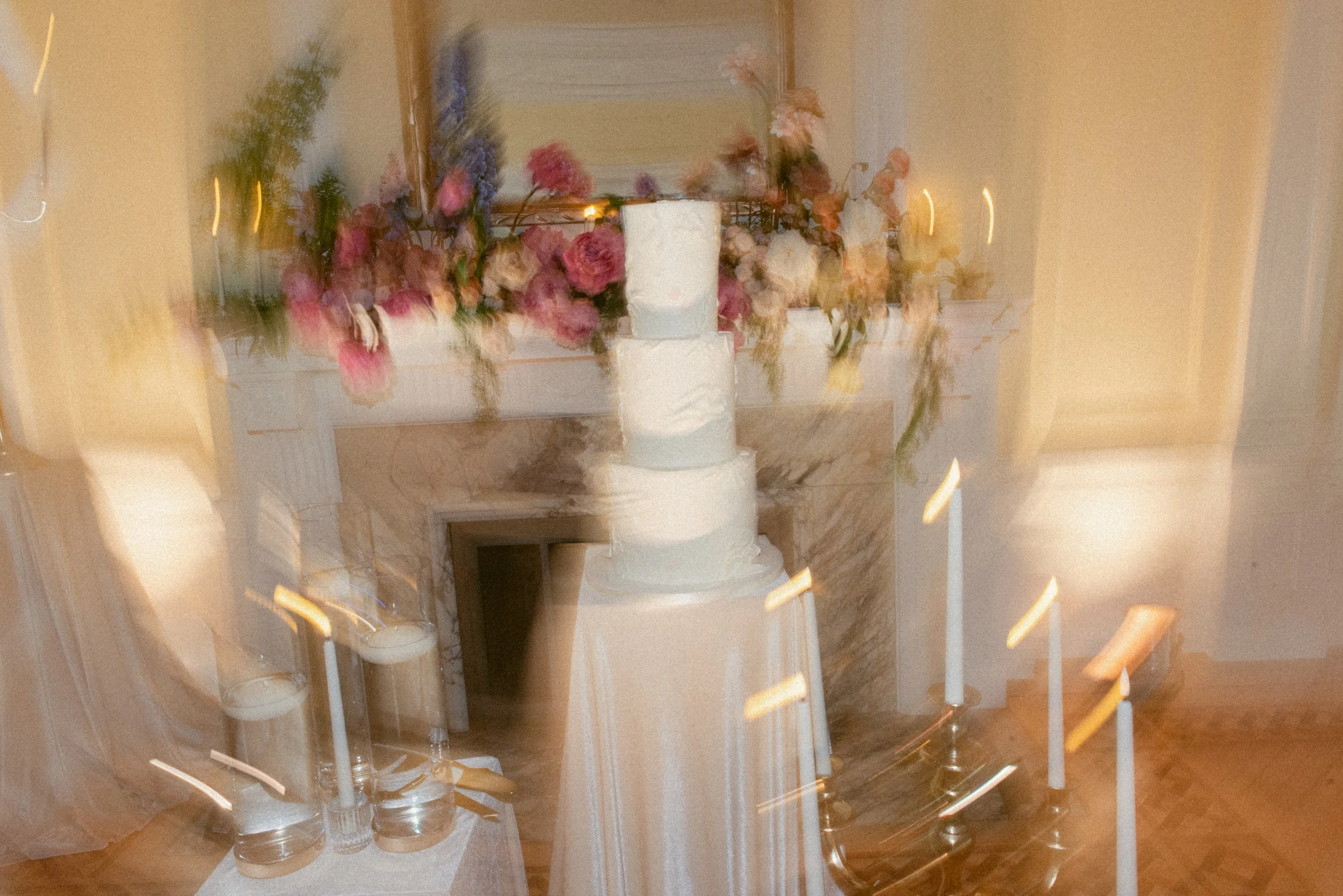 Wedding cake display at Cecil Green Park House, featuring a multi-tier white cake surrounded by glowing candles and soft motion blur, with pastel florals styled on the fireplace mantel.
