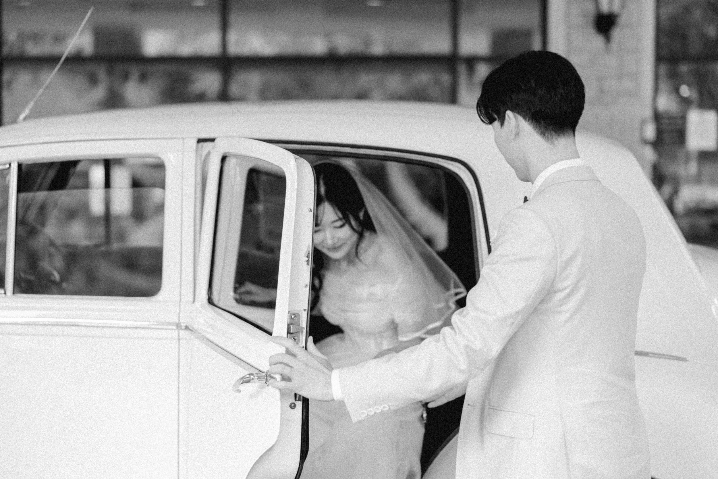 Black and white wedding photo of a groom opening the door of a vintage car as the bride steps out.