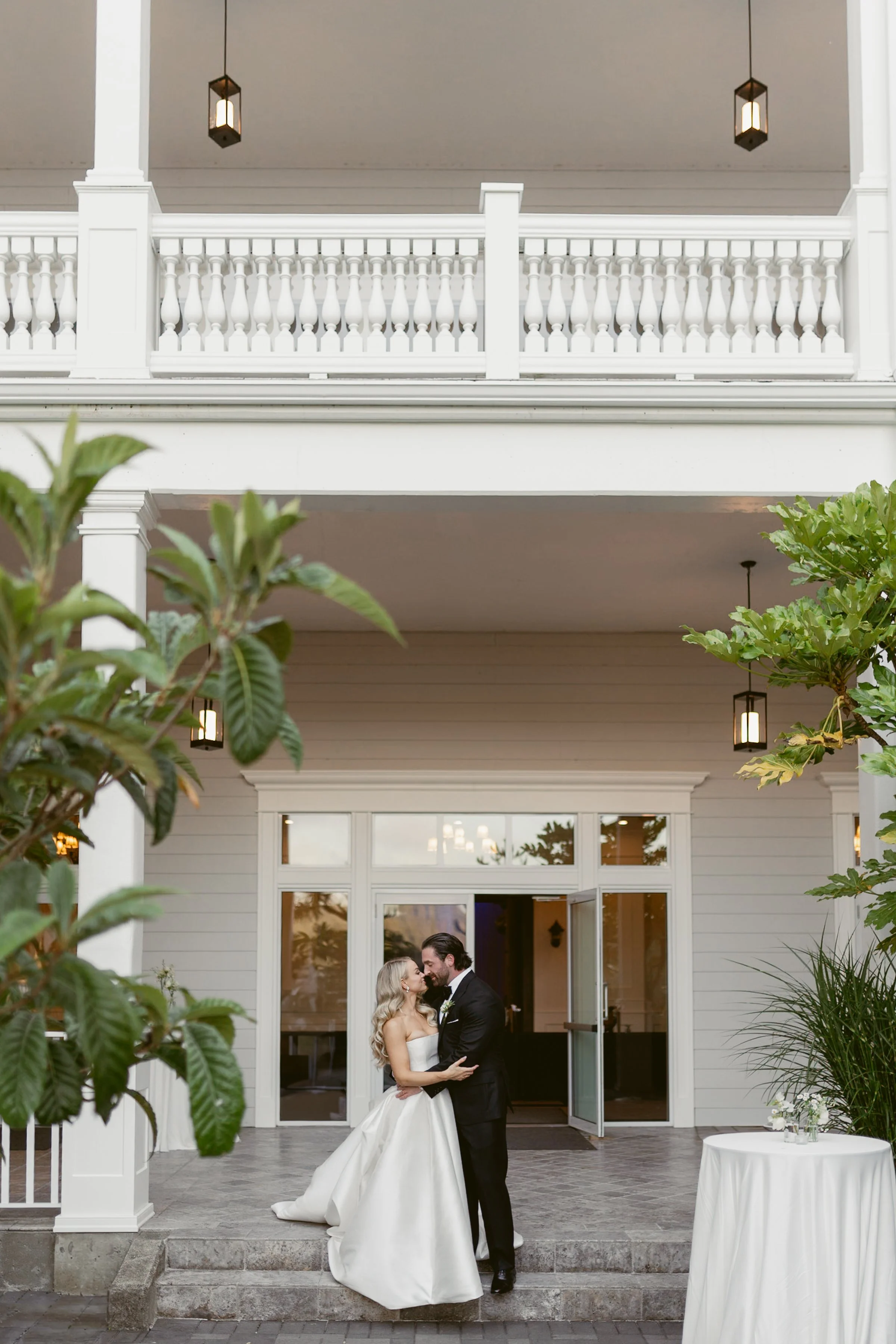 Bride and Groom touching noses on the veranda with greenery in the foreground.