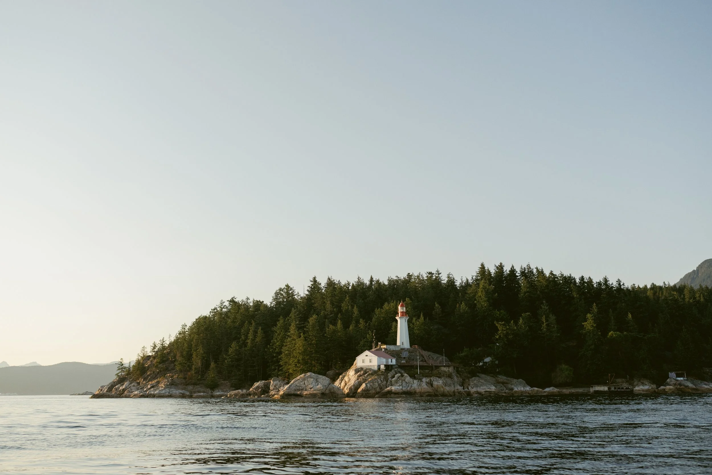 Coastal lighthouse surrounded by evergreen trees during golden hour, photographed from the water at a Vancouver yacht engagement party.