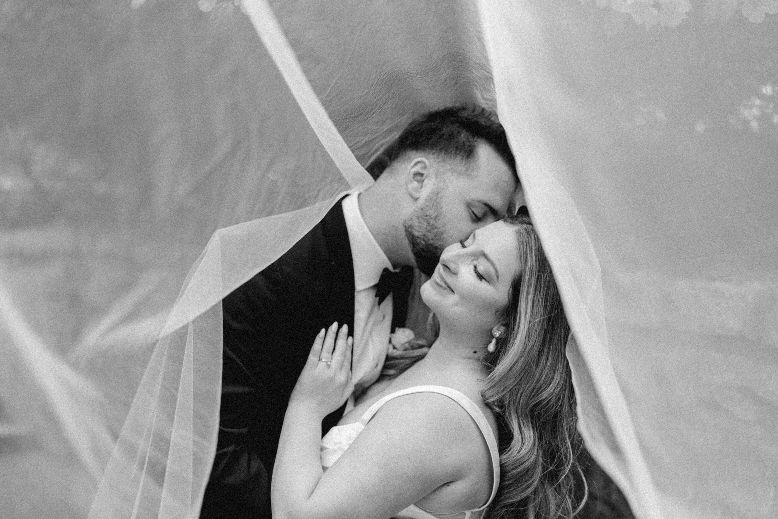 Black and white romantic wedding portrait at Queen Elizabeth Park, with the couple wrapped in a flowing veil as they share an intimate moment.