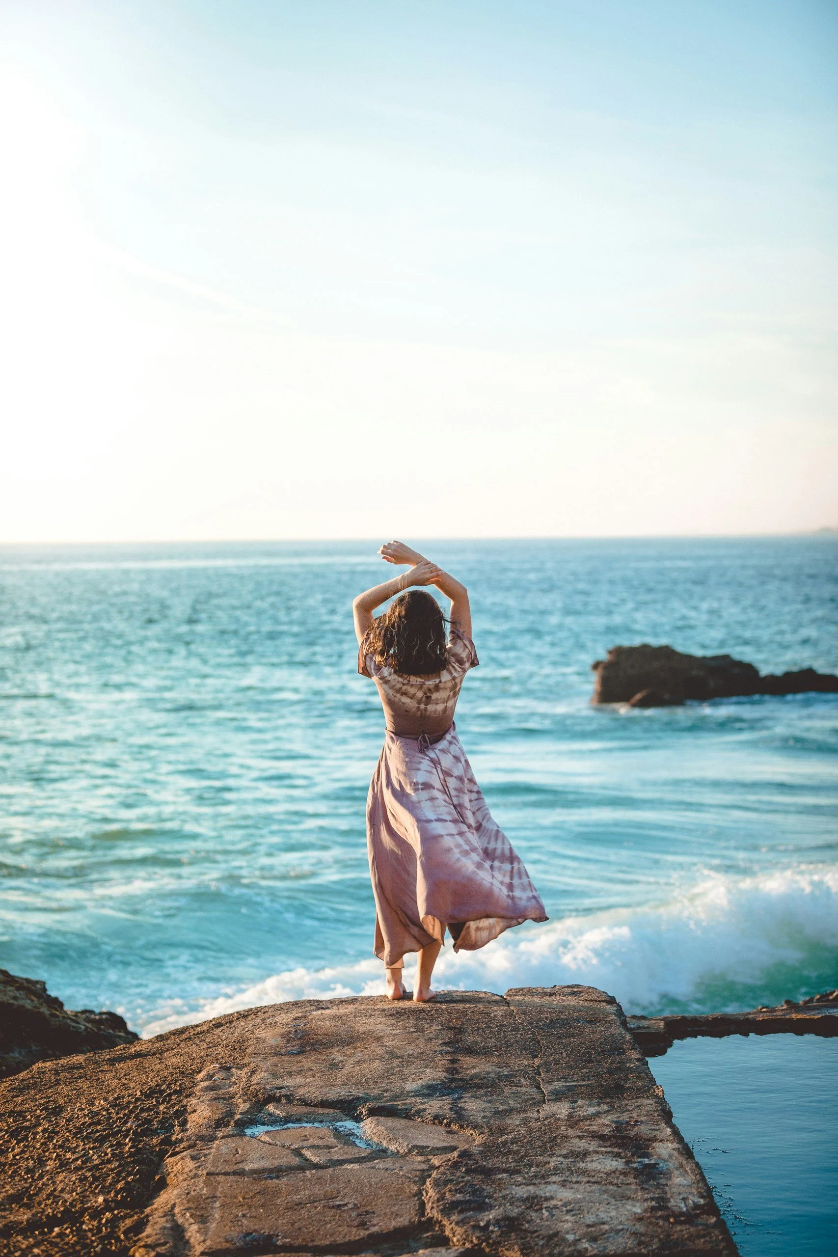 Woman standing on a rock with arms raised above her head looking out at the ocean representing empowerment and vitality in PMOS care