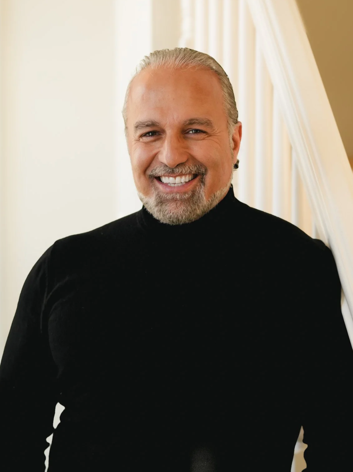 Smiling middle-aged man with silver hair and beard wearing a black turtleneck sweater, standing indoors in front of a cream-colored wall with curtains.