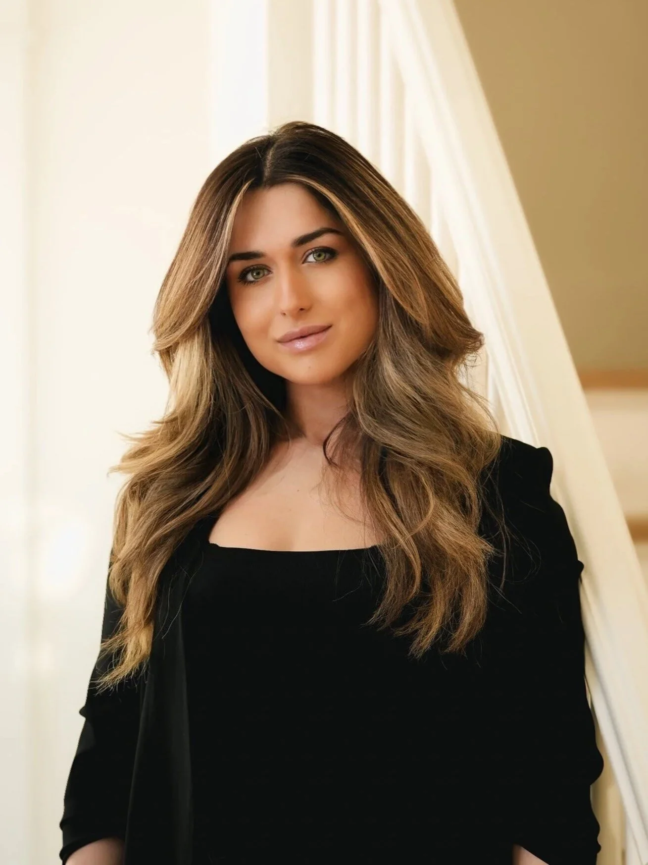 A woman with long, wavy, light brown hair and green eyes posing indoors beside a cream-colored curtain.