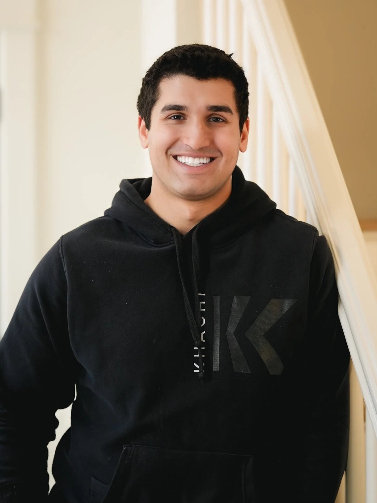 A smiling young man with short dark hair wearing a black hoodie, standing indoors near a staircase with white railings.