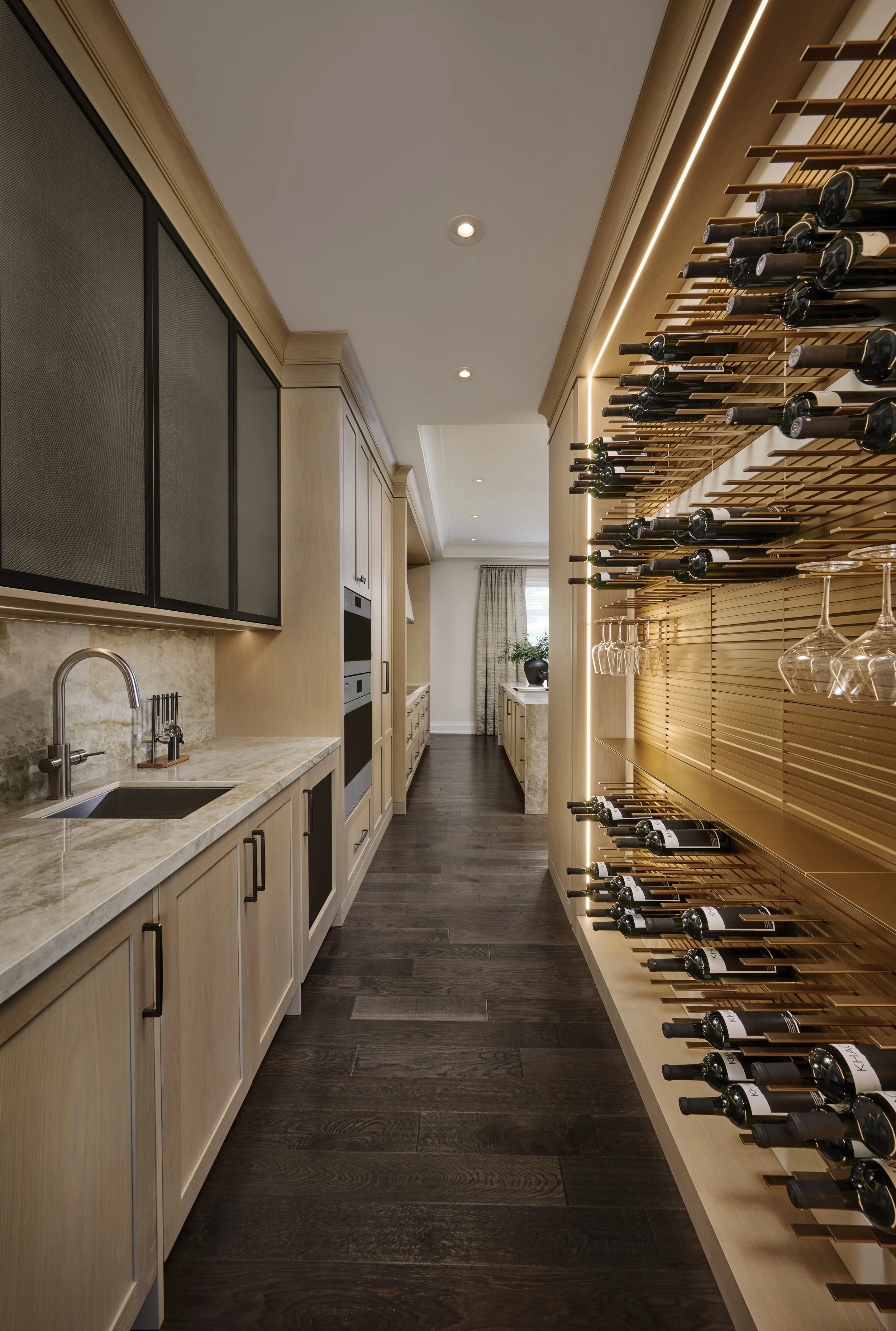 Modern kitchen with wooden cabinets, black hardware, marble countertop, and a wine rack with bottles on the right wall. There is a window with curtains at the end of the room, dark wooden flooring, and recessed lighting.