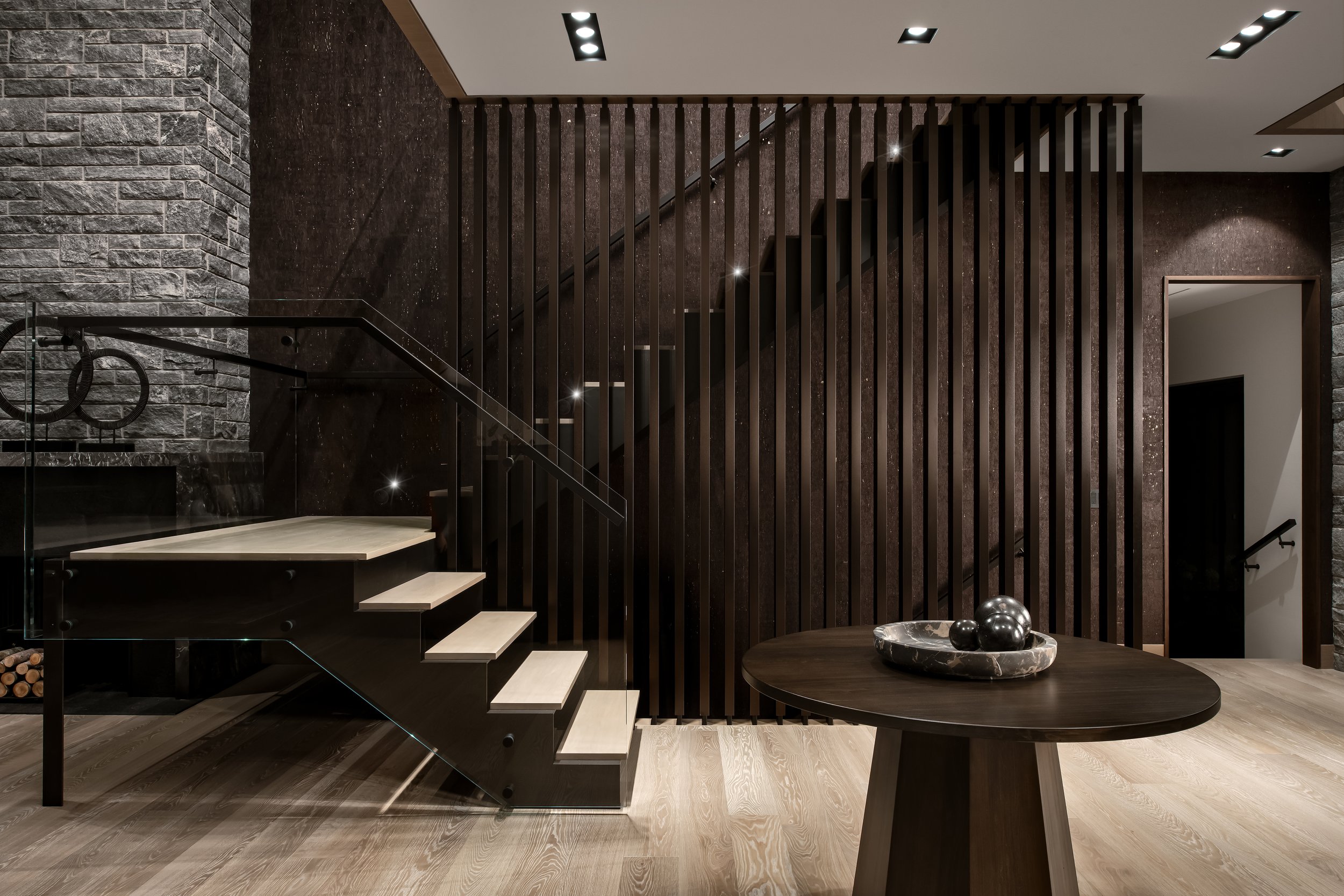 Modern interior with a staircase, stone wall, dark textured wall, wooden slats, and a round table with decorative spheres.
