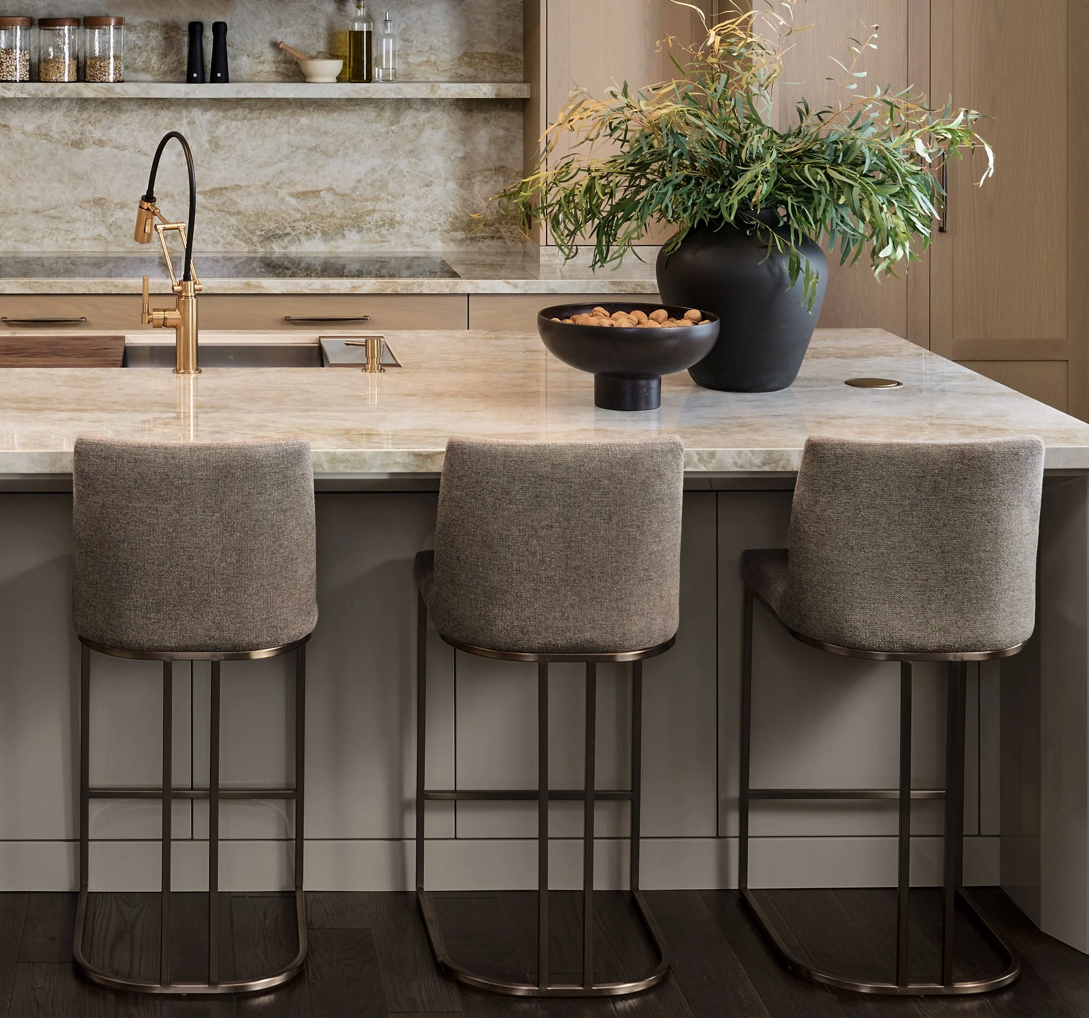 Modern kitchen with marble countertops, a gold and black faucet, large black vases containing greenery, and kitchen shelves with jars and bottles