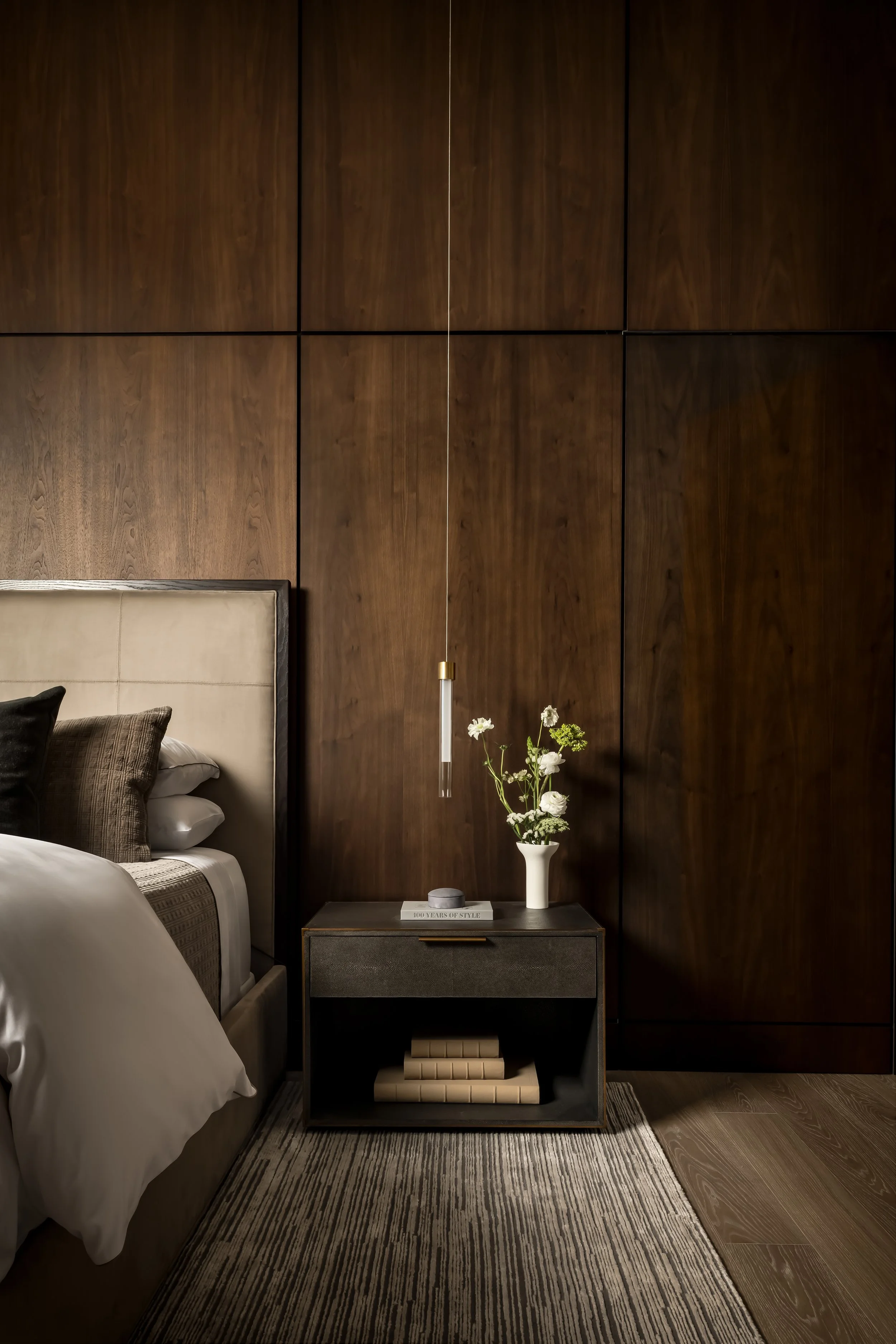 A bedroom corner with a bed on the left, a nightstand with a book and vase of flowers, dark wood paneled wall, striped rug, and hanging pendant light.