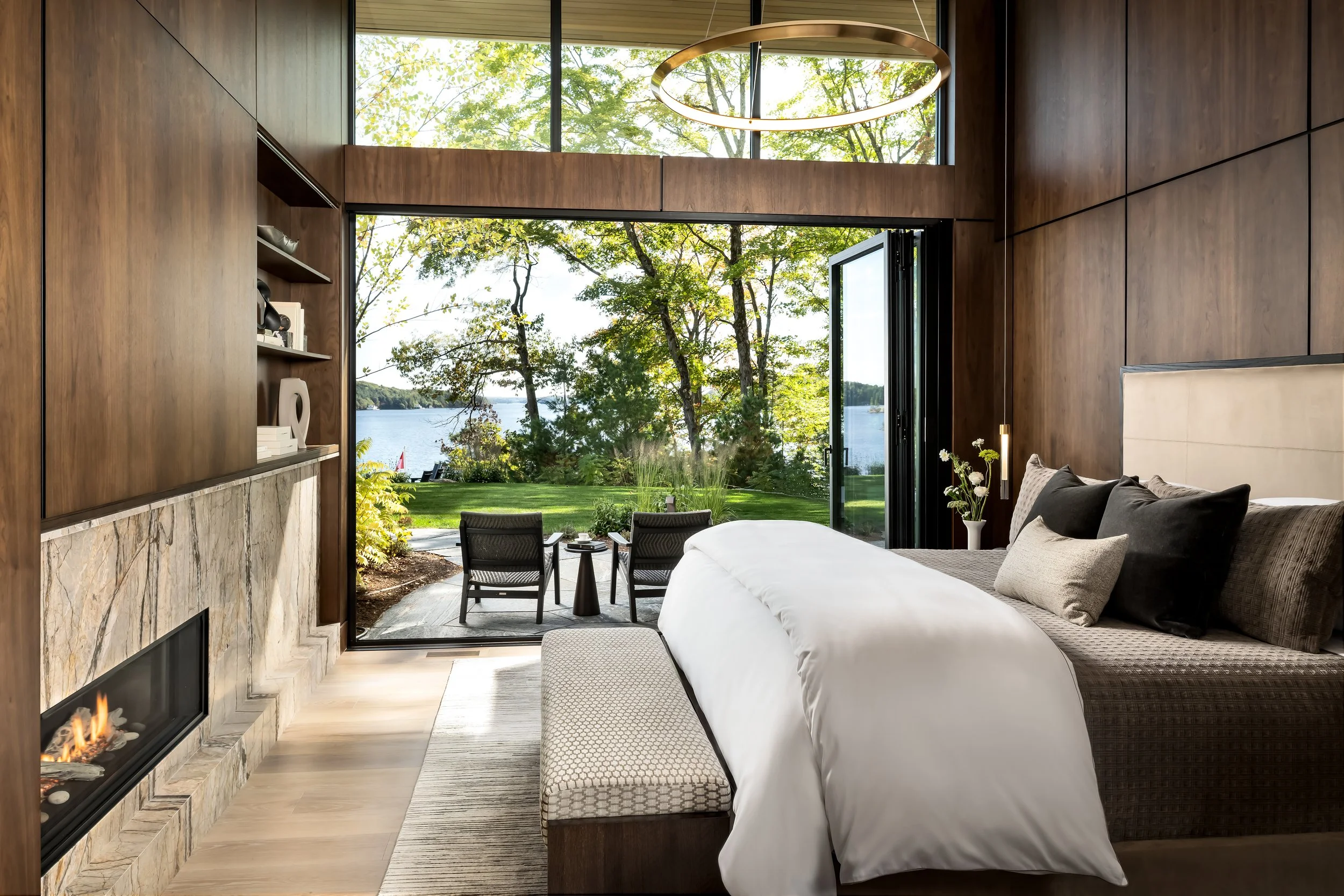 A modern bedroom with large windows looking out to a lakeside view. The room features wood-paneled walls, a white bed with pillows, a built-in fireplace, and a circular ceiling light fixture. Outside, there is a patio with chairs and a scenic landscape of trees and water.