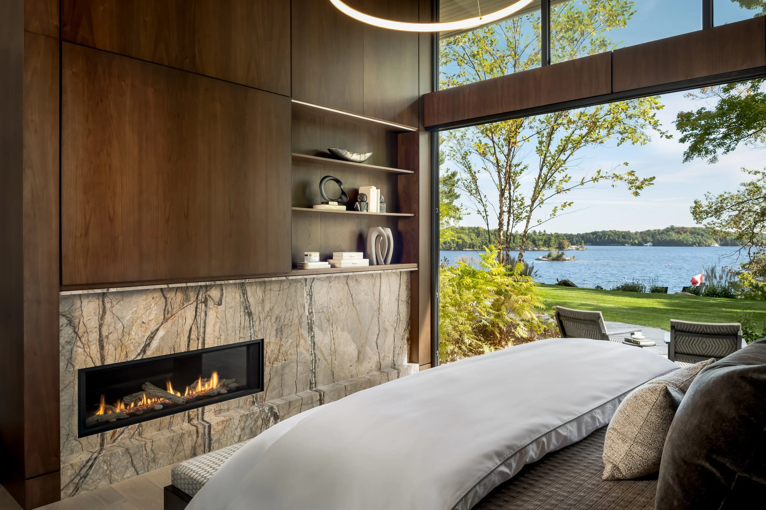 Modern bedroom with a large floor-to-ceiling window showing a lakeside view, featuring trees, outdoor chairs, and a small flag by the water, with a fireplace and wooden shelves inside.