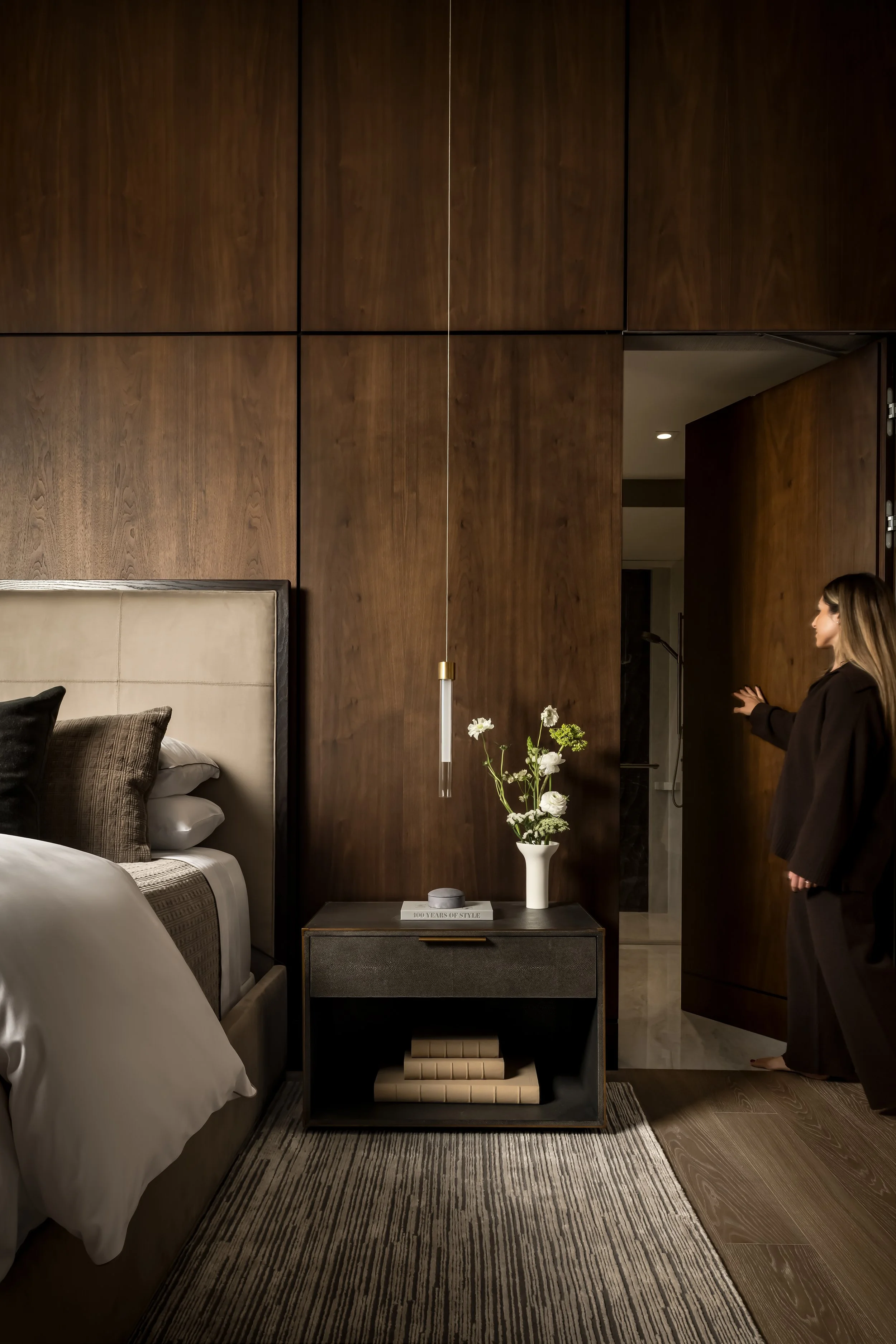 A bedroom with a beige upholstered bed, a dark wooden nightstand with a white vase holding flowers, and a tall hanging pendant light. There is a woman with long hair and a black blazer standing near a doorway, talking or signaling. The room has dark wood-paneled walls and a patterned area rug.