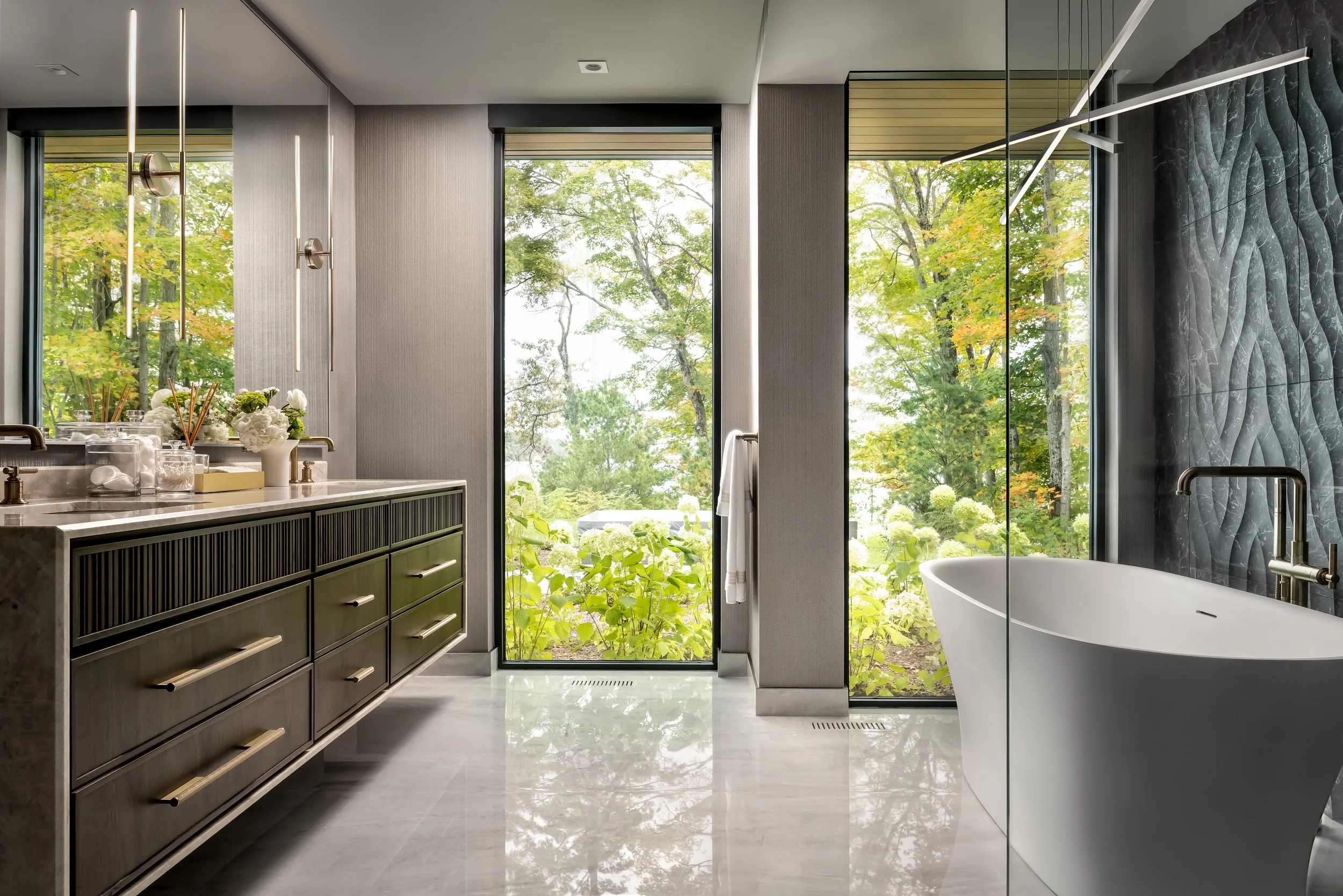 Modern bathroom with large windows showing green trees outside. Features a white bathtub, a dark green cabinet with gold handles, and decorative flowers on a marble countertop. Wall has textured gray tiles, and ceiling has two minimalist vertical light fixtures.