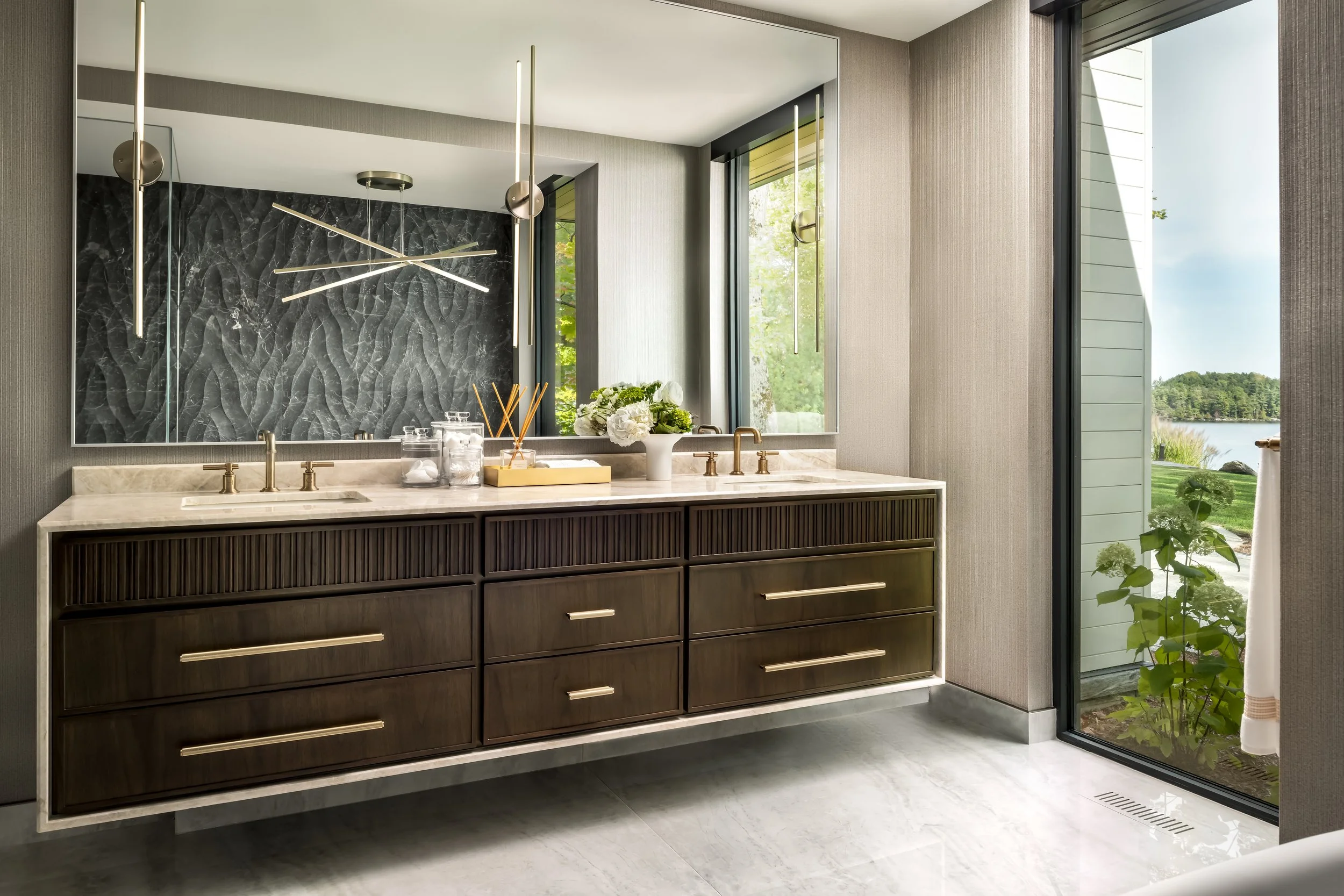 Modern bathroom with dark wood vanity, marble countertop, gold fixtures, large mirror, and large windows with a view of trees and water.