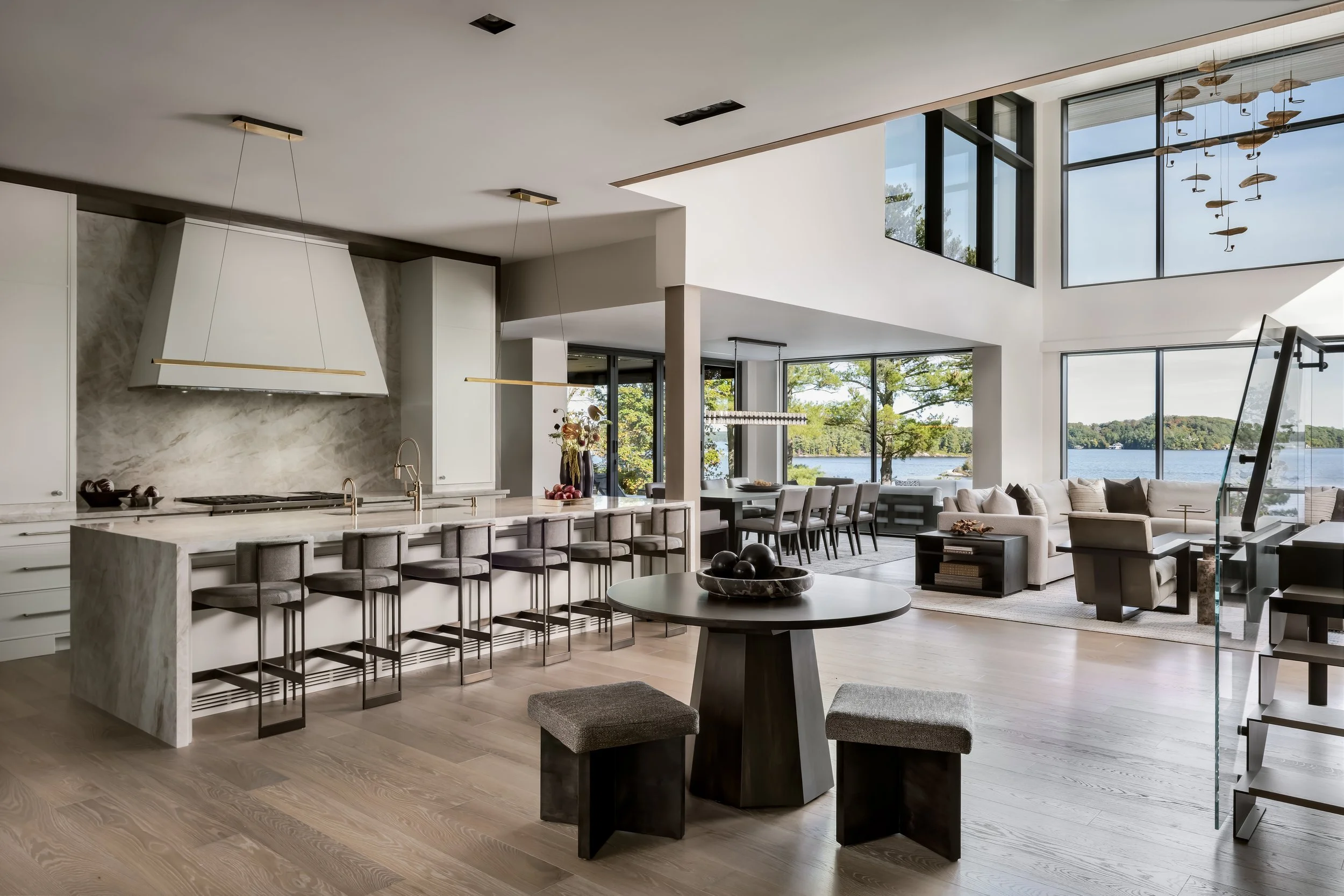 Modern, open-concept living space with large windows overlooking a lake, featuring a kitchen with white cabinetry and marble countertops, a dining area with a long table and chairs, and a cozy seating area with couches and a TV, decorated with minimalist furniture and decor.