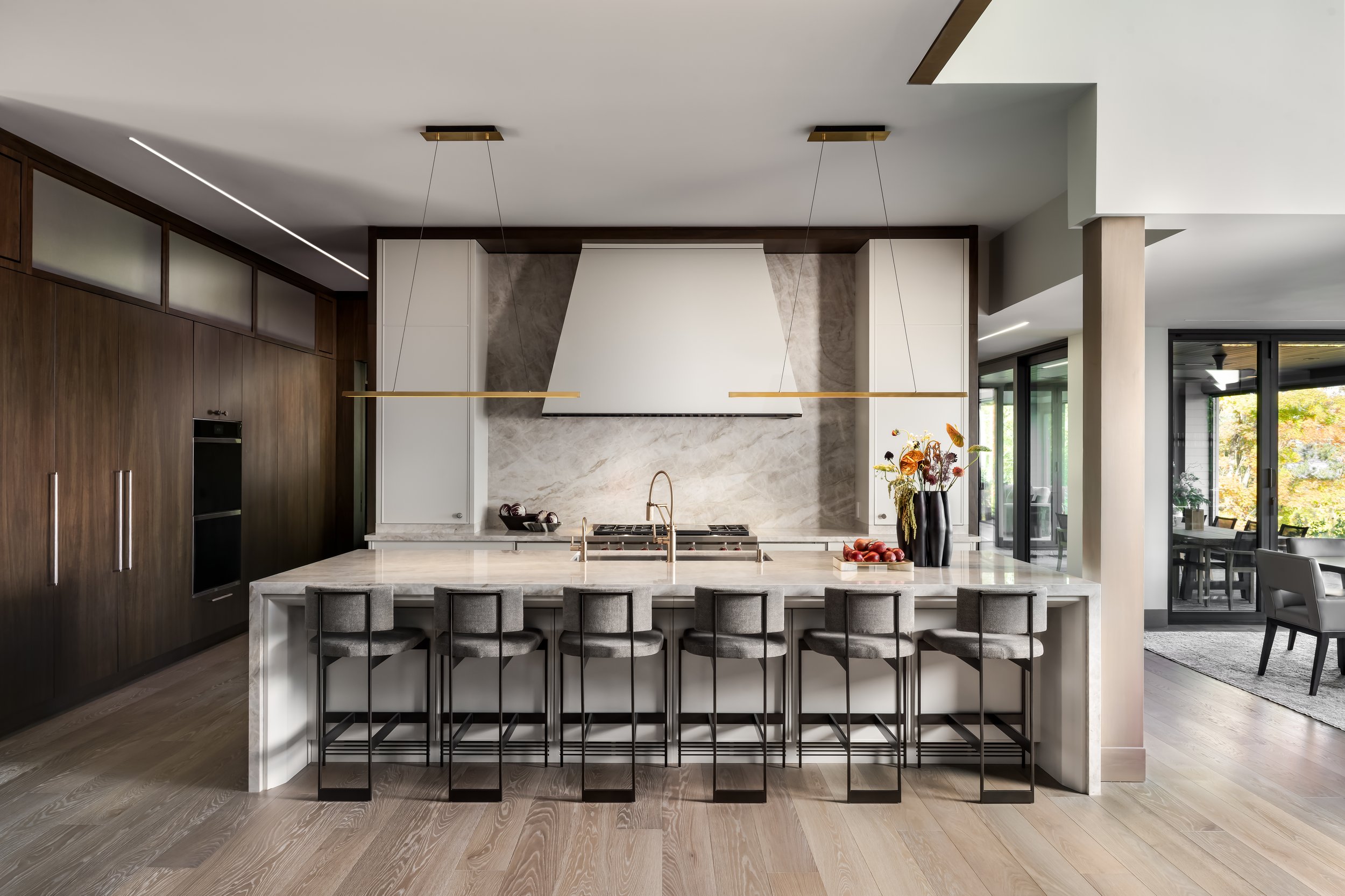 Modern kitchen with a large marble island, dark wood cabinets, white wall and a window with a view of trees outside.