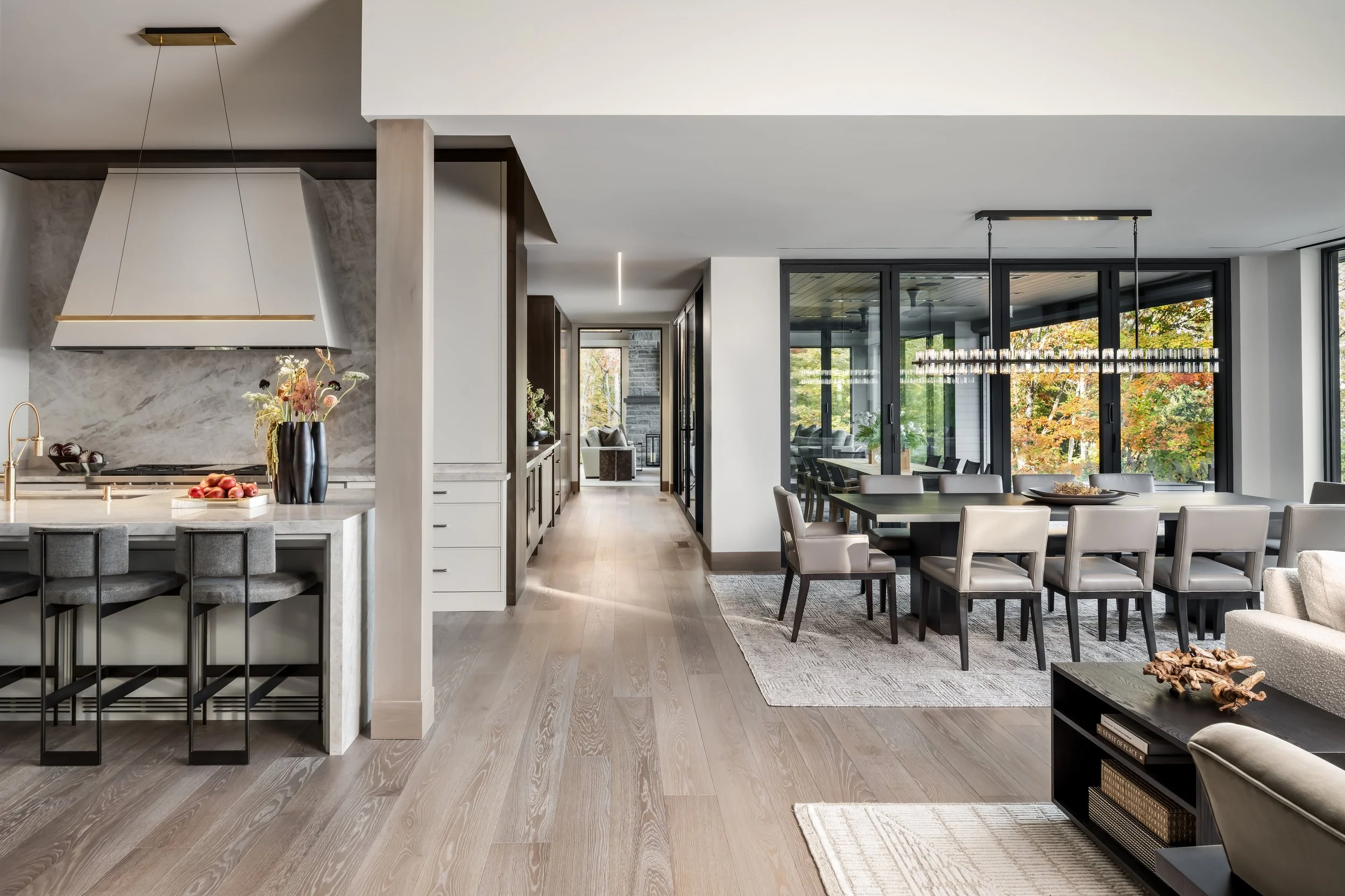 Open-concept modern living space with kitchen and dining areas, large windows with view of trees, and neutral-colored furniture.