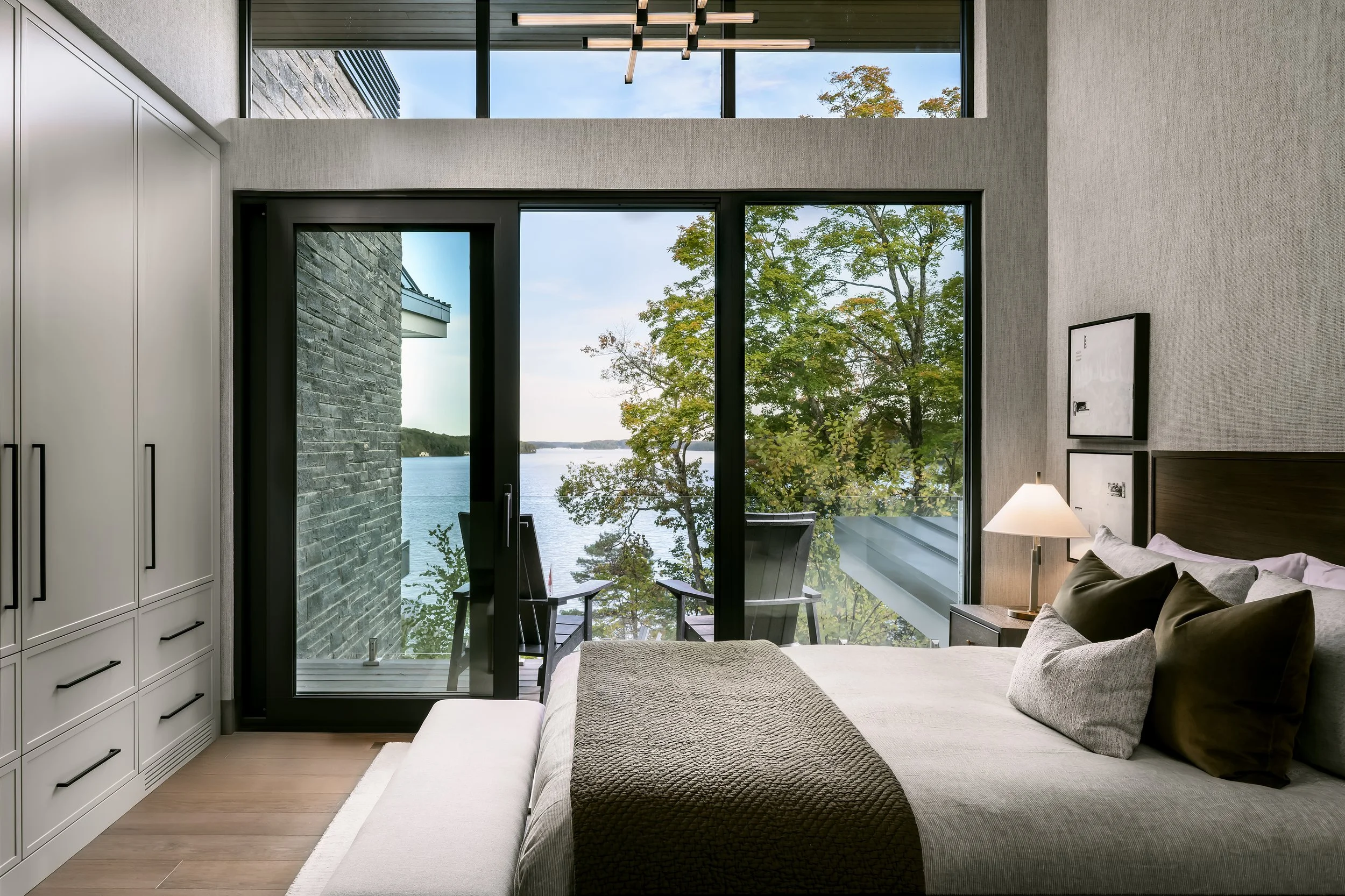 Modern bedroom with large glass windows and a view of trees and water outside. The room has a white wardrobe, a bed with gray and white bedding, a bedside table with a lamp, and outdoor seating on a small balcony.
