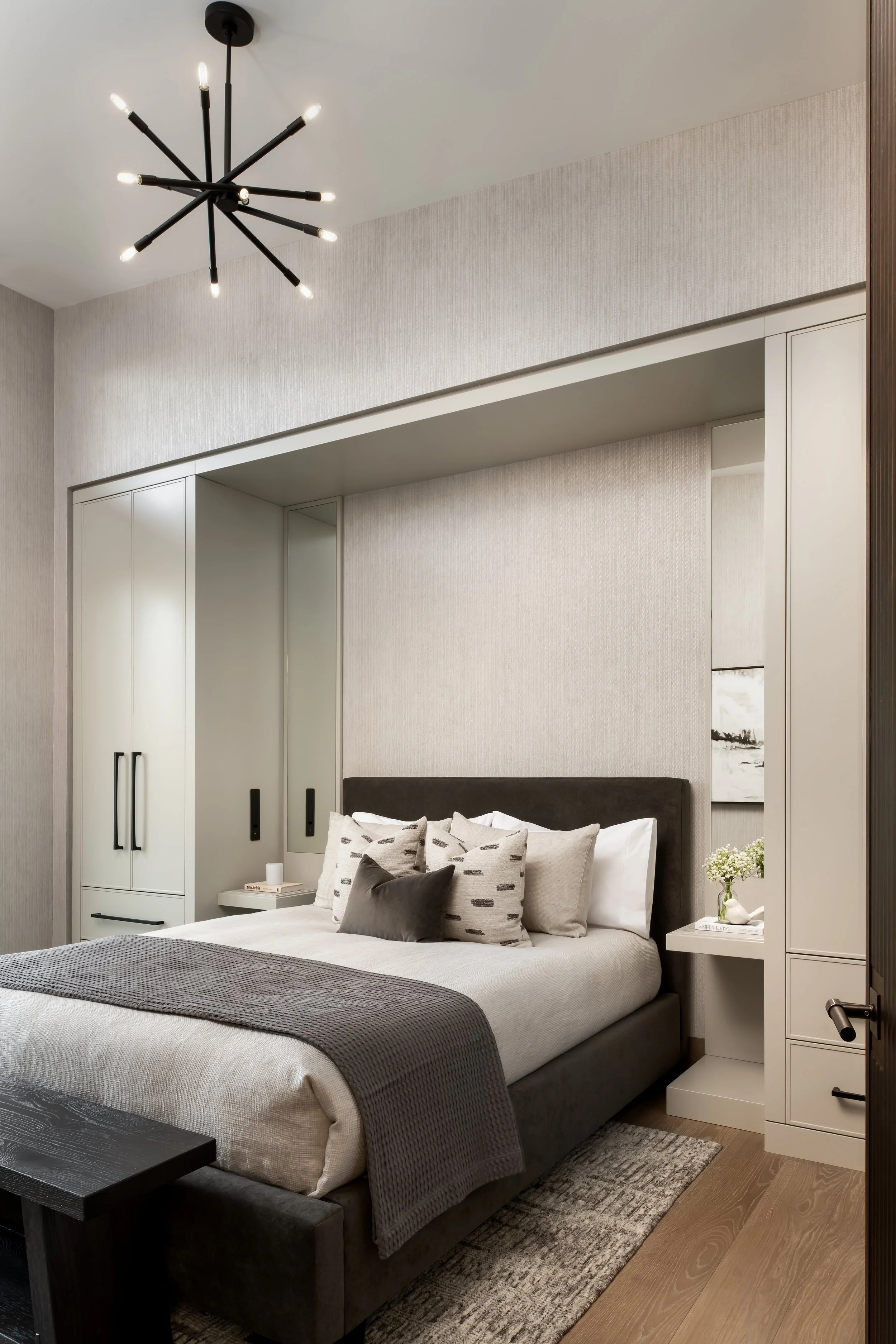 Modern bedroom with a bed featuring multiple pillows, a dark headboard, and white bedding. White built-in cabinets and a bedside table with a small bouquet of flowers and books. A contemporary black chandelier hangs from the ceiling, and a textured grey and beige rug covers part of the wooden floor.