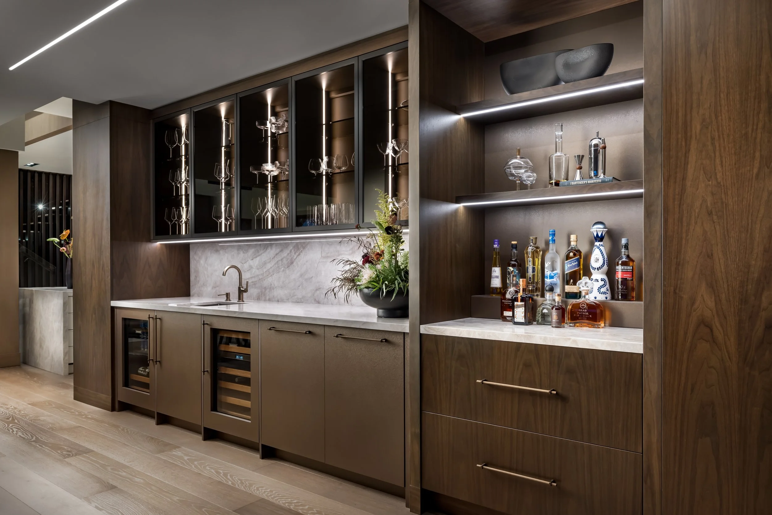 Modern kitchen with dark wood cabinets, a marble countertop, and a glass-front wine fridge, decorated with a vase of flowers and a bar area with various bottles and glassware.