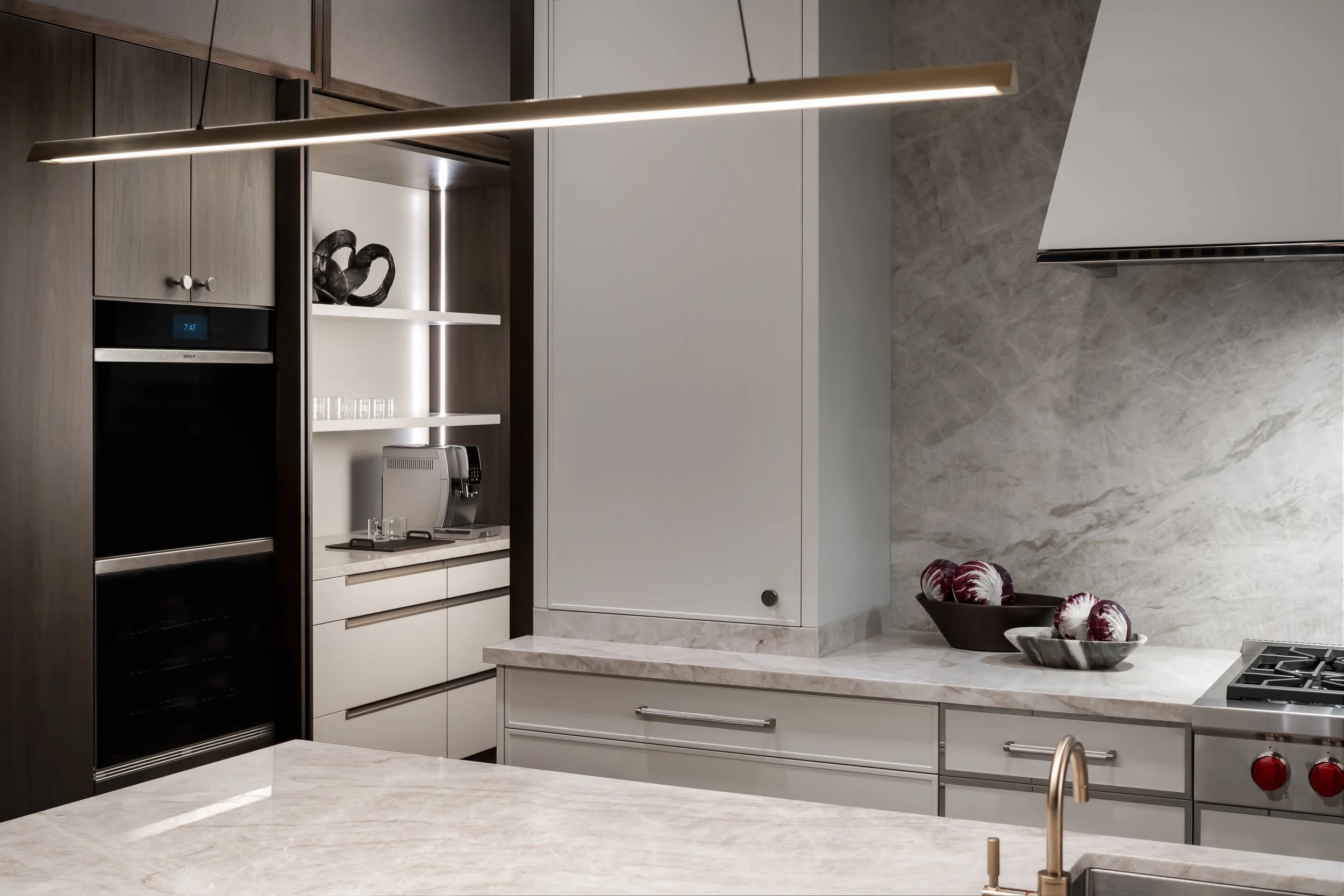 Modern kitchen with marble countertops, a black oven, white cabinets, a decorative sculpture on a shelf, a bowl of radicchio, and a sleek overhead light fixture.