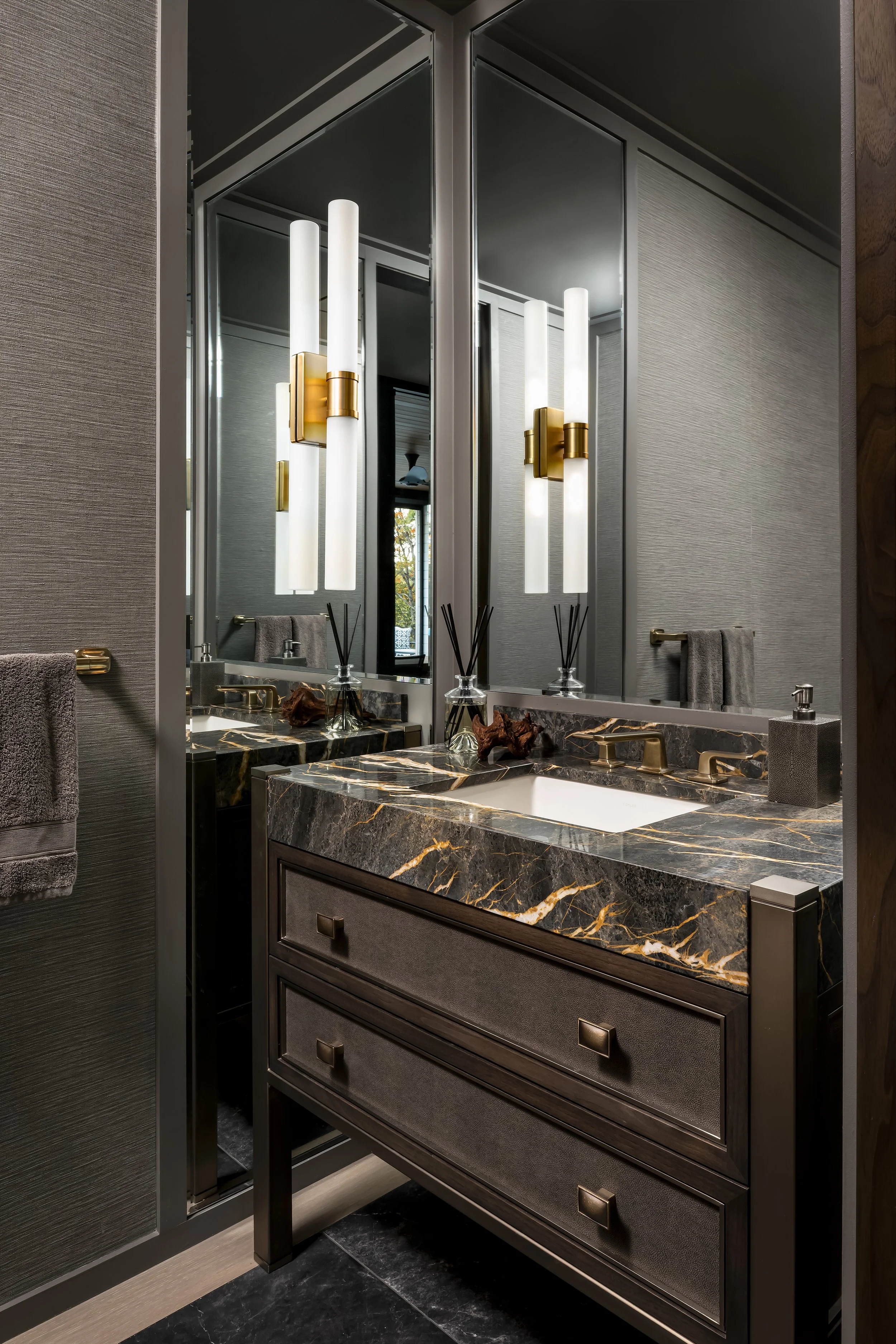 A modern bathroom vanity with a dark-colored marble countertop, gold fixtures, and a large mirror. There are wall-mounted lights with elongated white shades and a towel hanging on a hook to the side.