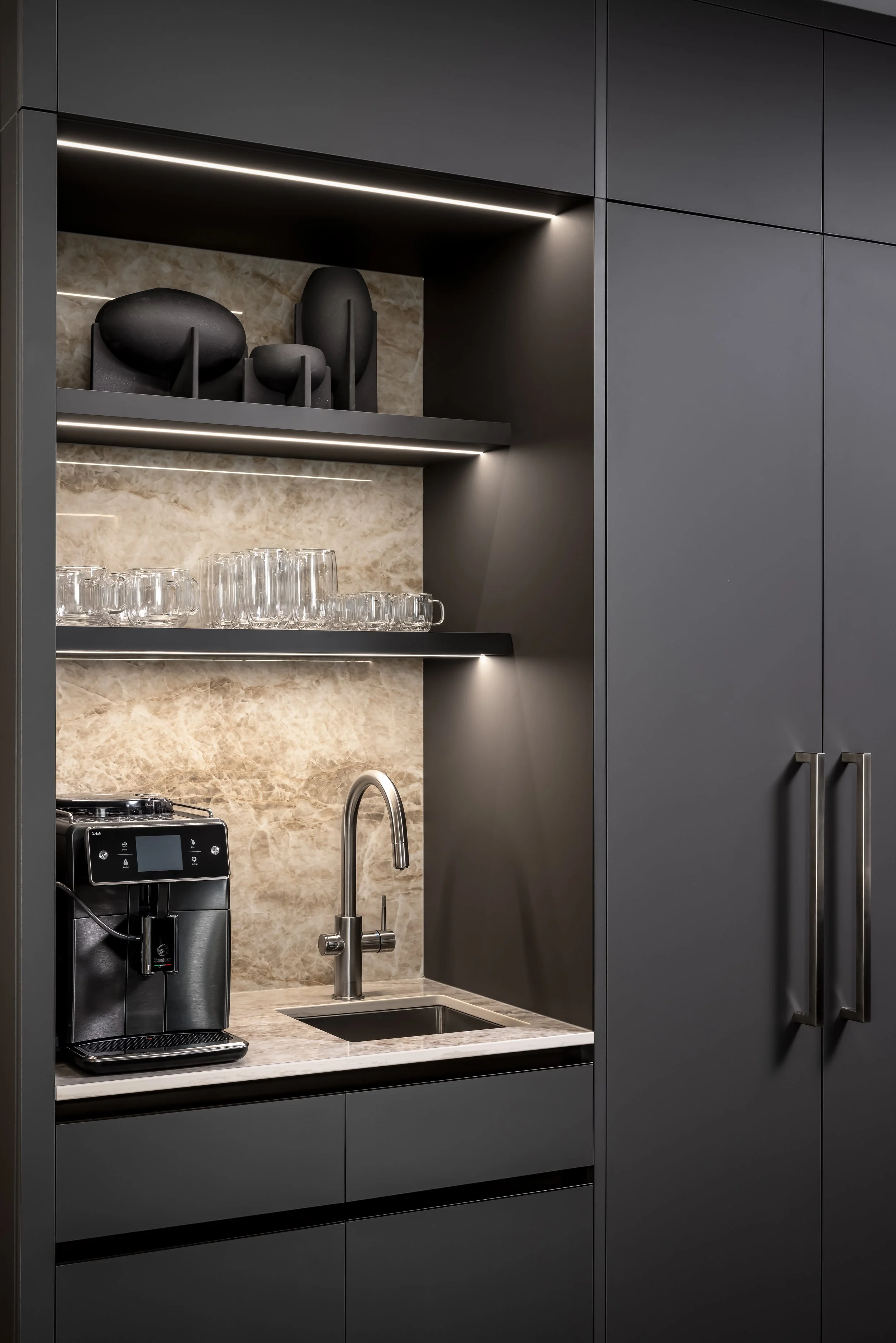 Modern kitchen with dark gray cabinets, a beige marble backsplash, and built-in shelves holding black decorative objects and glassware. There is a small stainless steel sink, a faucet, and a coffee machine on the countertop.